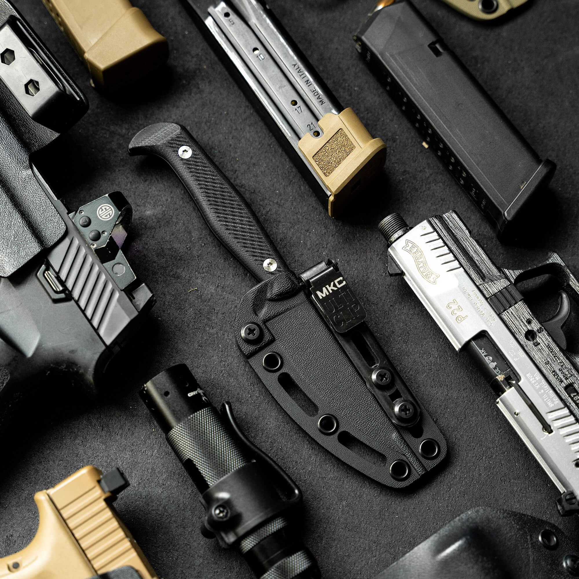 Alt text: "Close-up view of a tactical black Battle Goat knife with textured handle, displayed among various military equipment including magazines, firearm parts, and other tactical gear on a dark background."