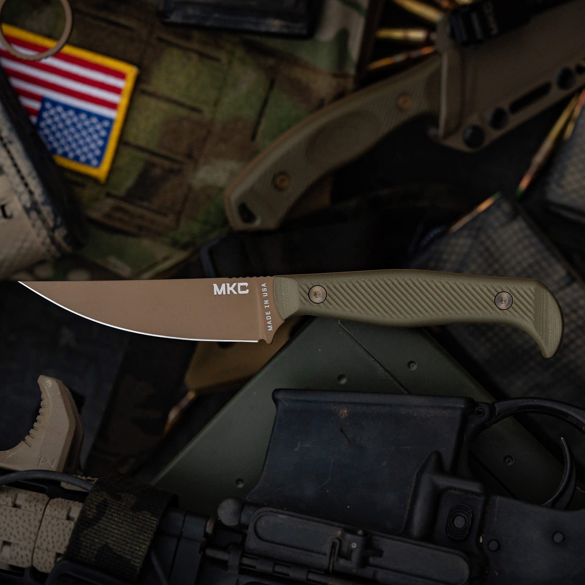 Close-up image of a battle goat folding knife from MKC in a muted coyote olive drab color, displayed on assorted tactical gear including a camouflage backpack, magazines, and parts of firearms.