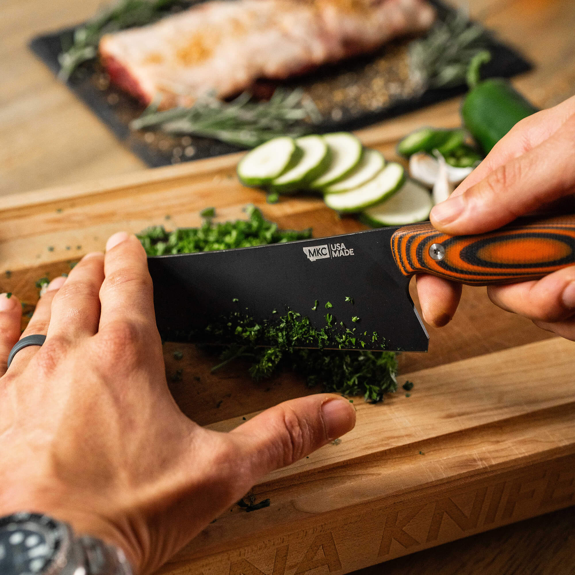 Person using a Bighorn Chef knife with an orange and black handle to chop fresh herbs on a wooden cutting board, with ingredients and prepared food visible in the background.