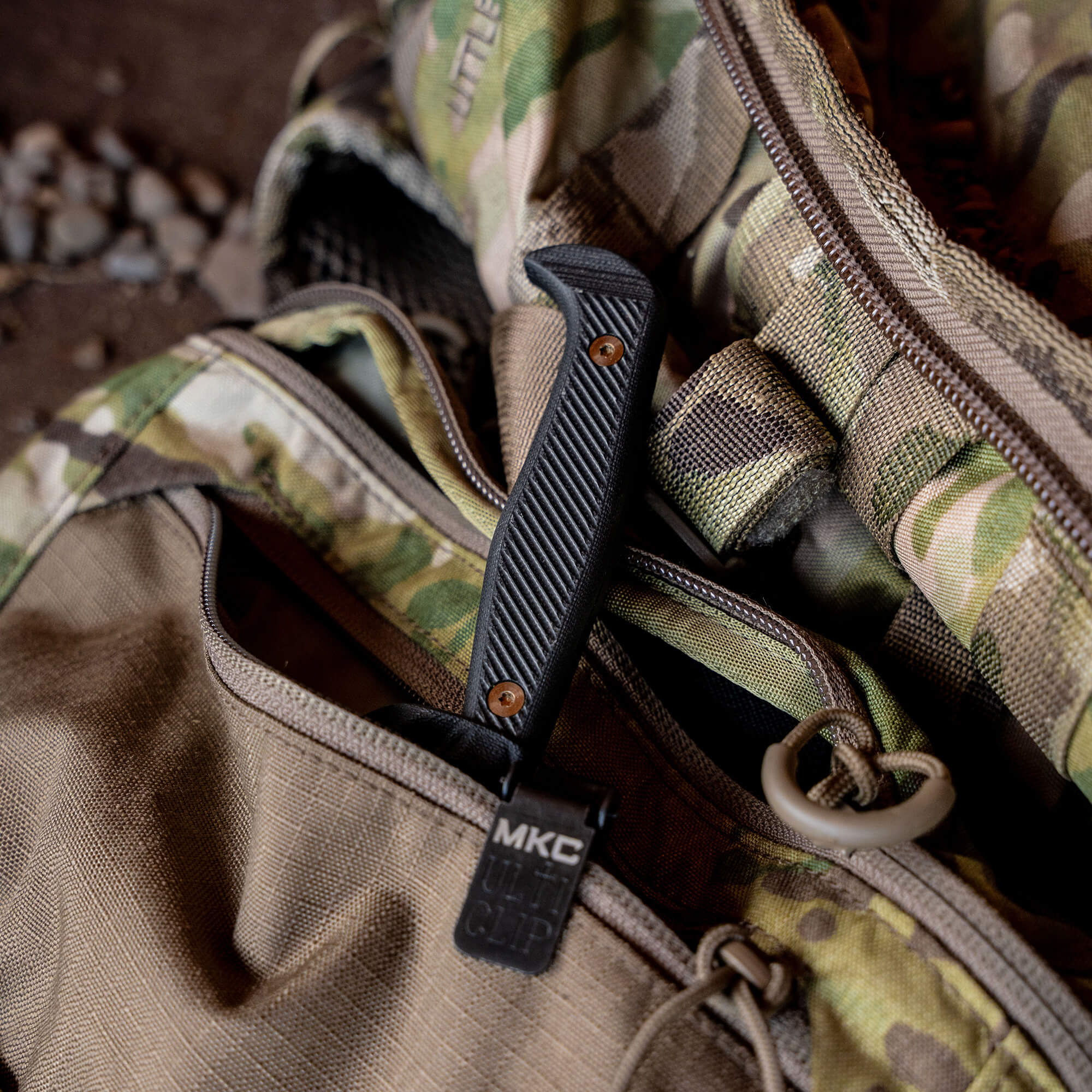 Close-up image of a black textured handle knife with an orange rivet and MKC tag, partially tucked into a camouflage military-style bag.