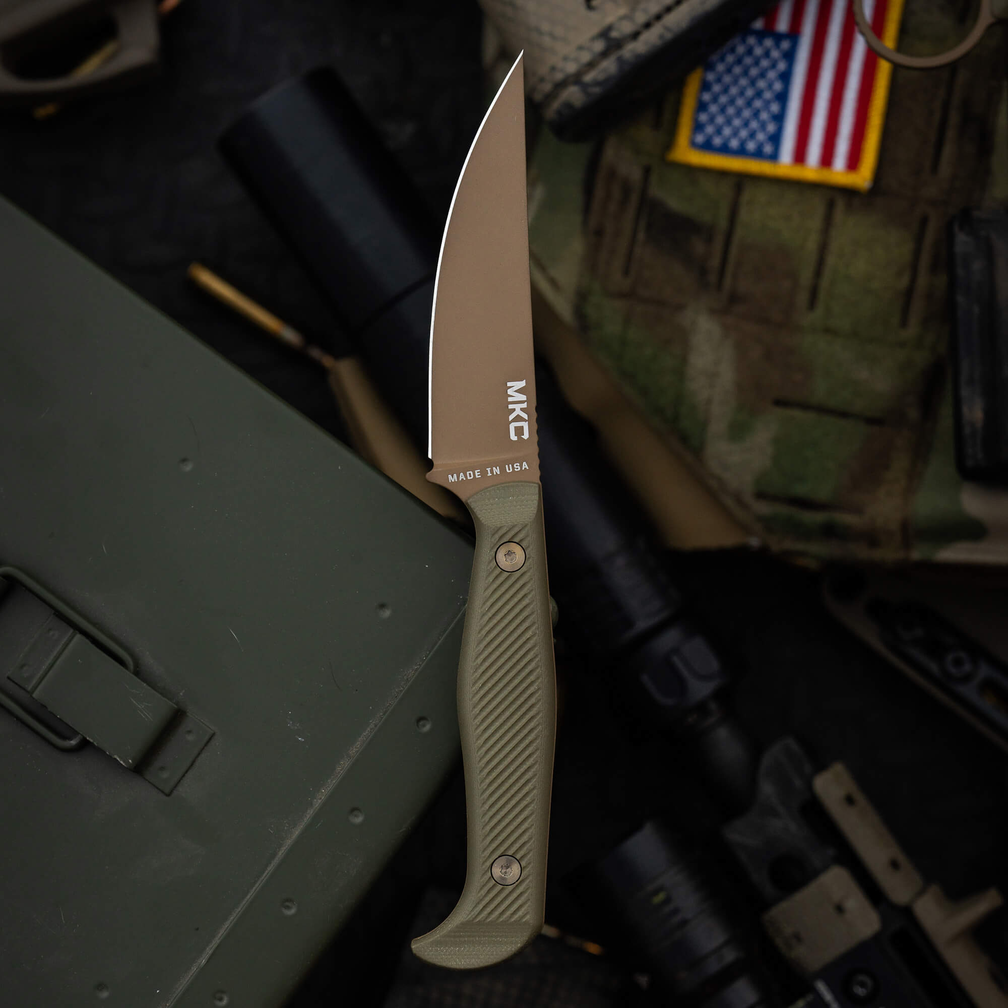 Alt text: "BATTLE GOAT tactical knife by MKC with a tan blade and olive drab handle, displayed against a military gear background, including an American flag patch. Made in USA inscription visible on the blade."