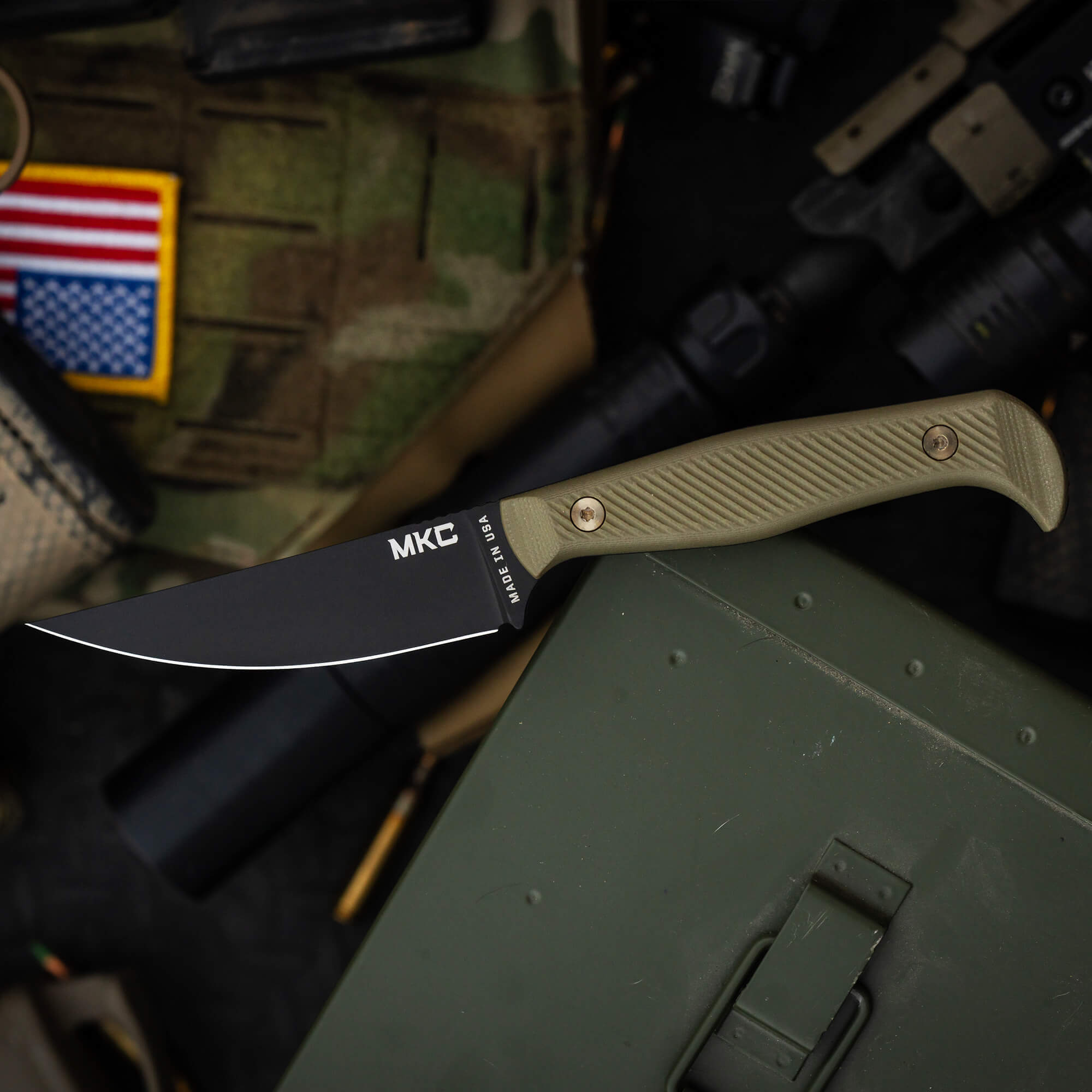 Alt text: A fixed-blade BATTLE GOAT MKC knife with a black blade and olive drab handle, resting on a green military box with tactical equipment and an American flag patch in the background.