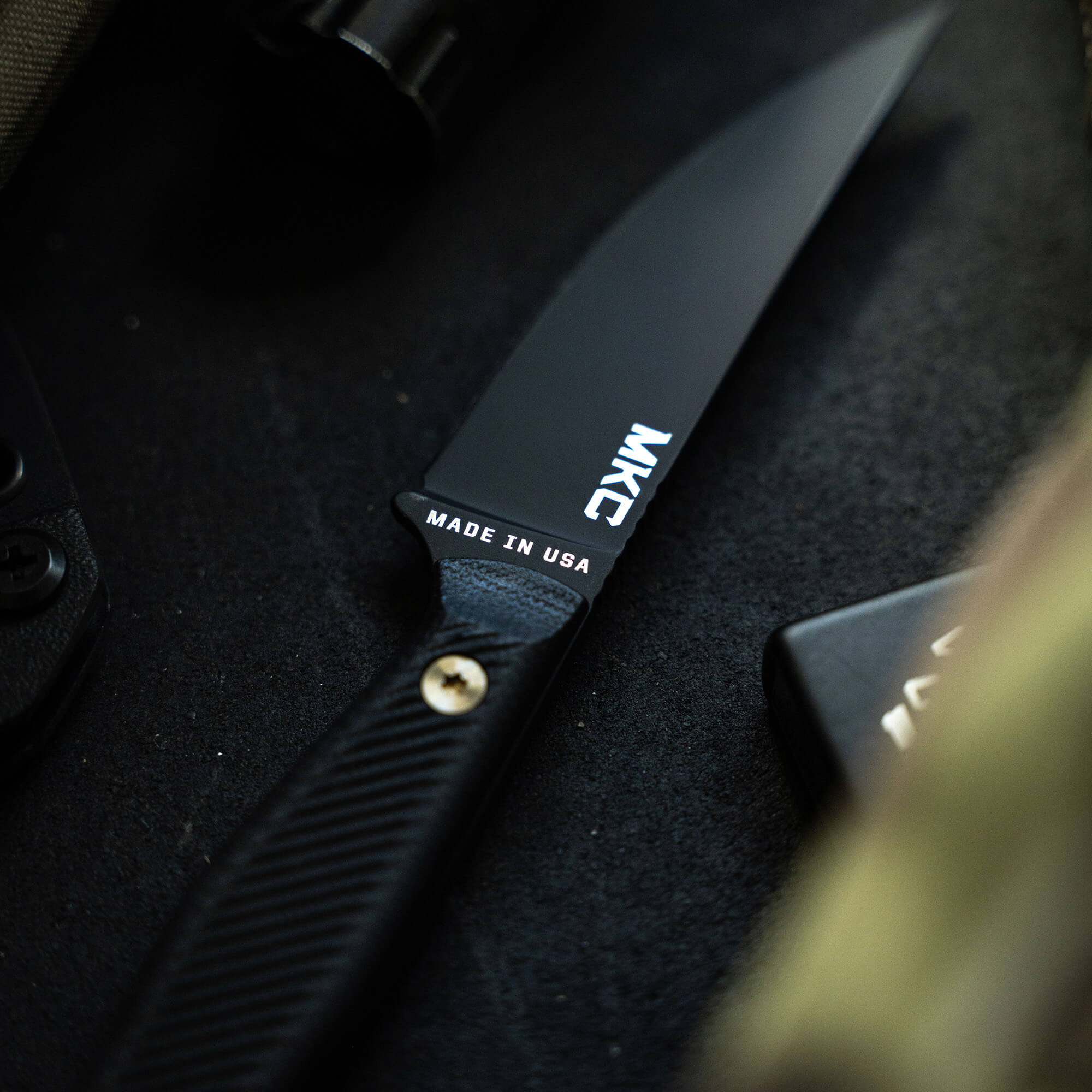 Close-up image of the Battle Goat knife in black, featuring a sleek blade with 'MKC' and 'MADE IN USA' printed on it, and a textured handle with visible rivets.