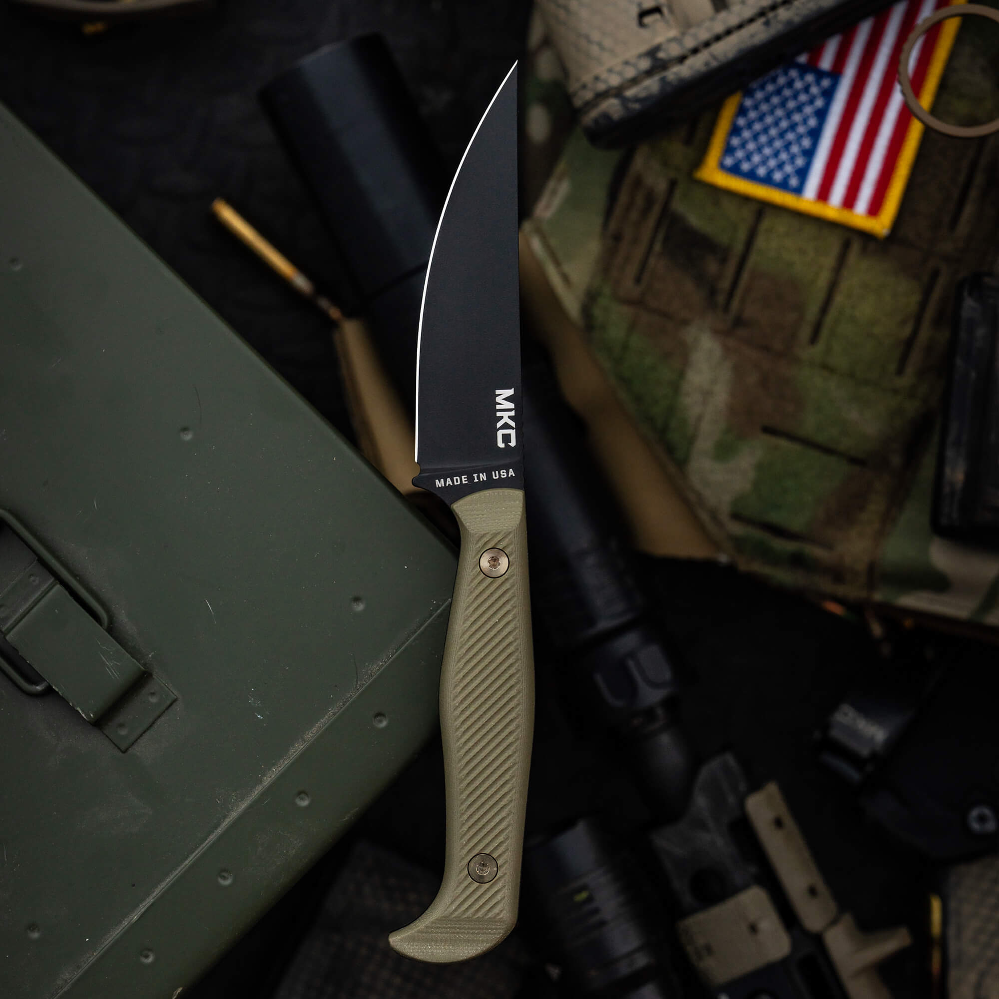 Alt Text: "Close-up of a MKC Battle Goat tactical knife with a black blade and olive drab handle, prominently featuring the text 'MADE IN USA'. The knife is displayed on a military-themed backdrop with a green ammo box, tactical
