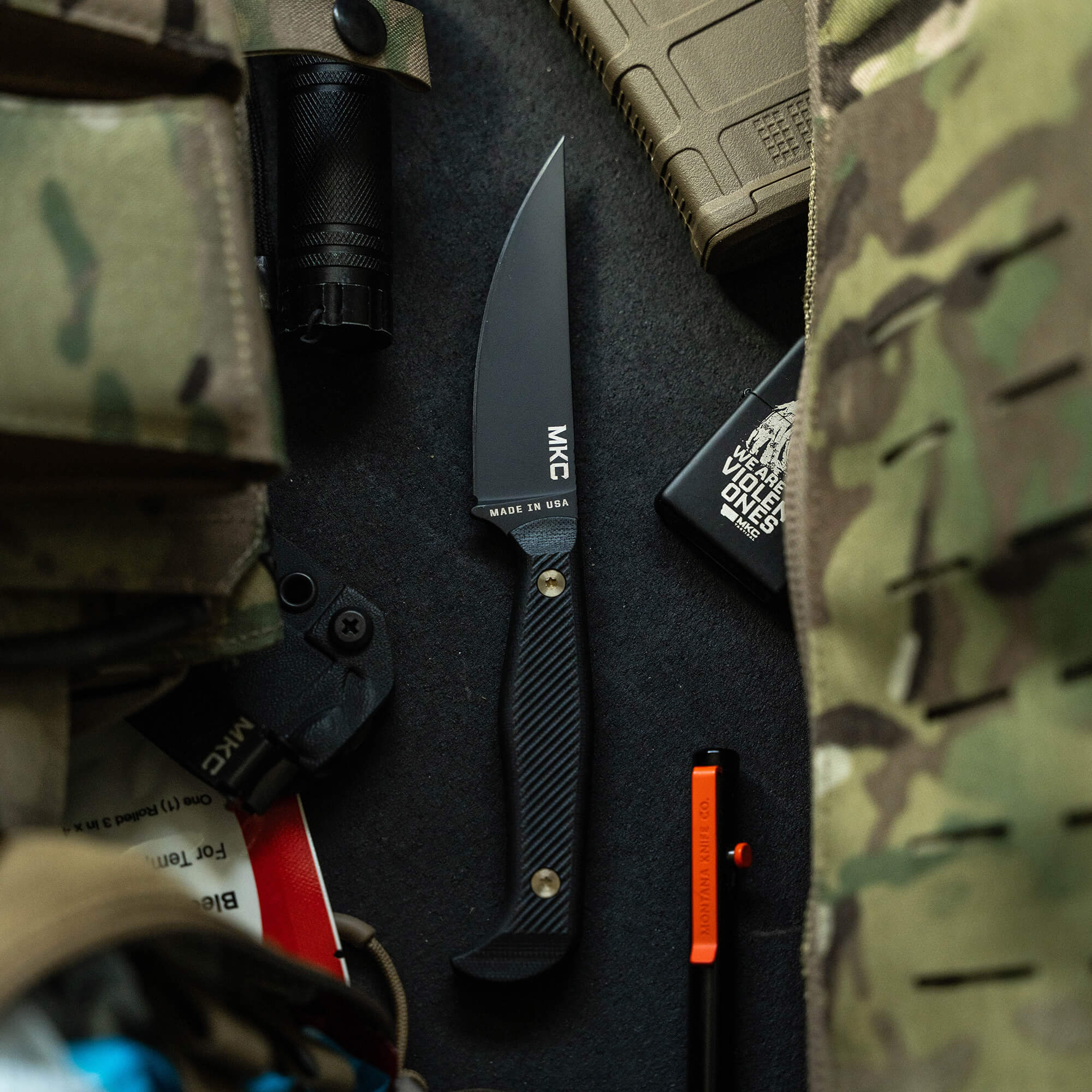 Alt text: "Black MKC Battle Goat tactical knife displayed on a tactical gear background, featuring a curved blade and carbon fiber handle, with 'MADE IN USA' text on the blade."