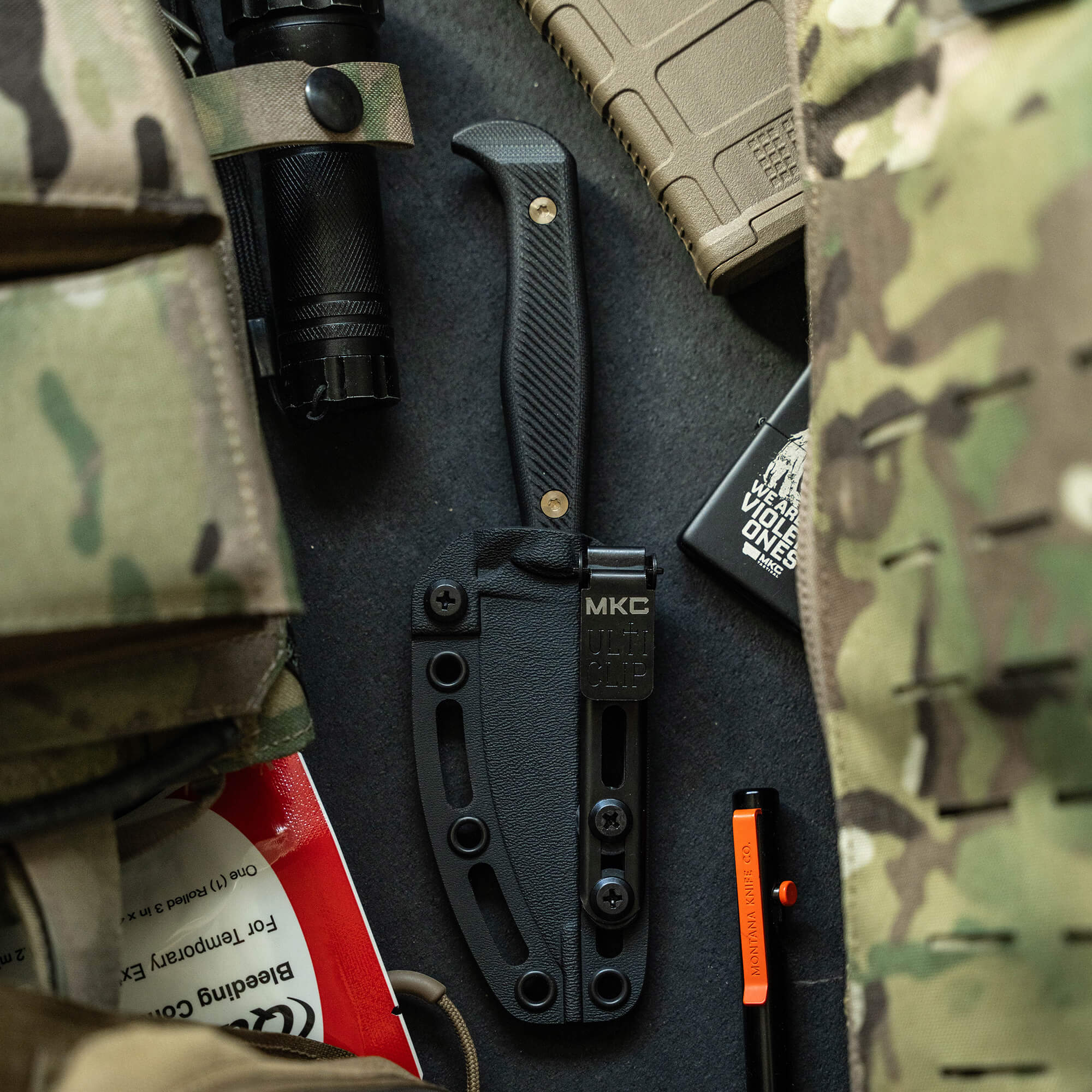 Alt text: "Close-up view of a black tactical knife, BATTLE GOAT - BLK/BLK, in a sheath attached to tactical gear including a flashlight and camouflage material."