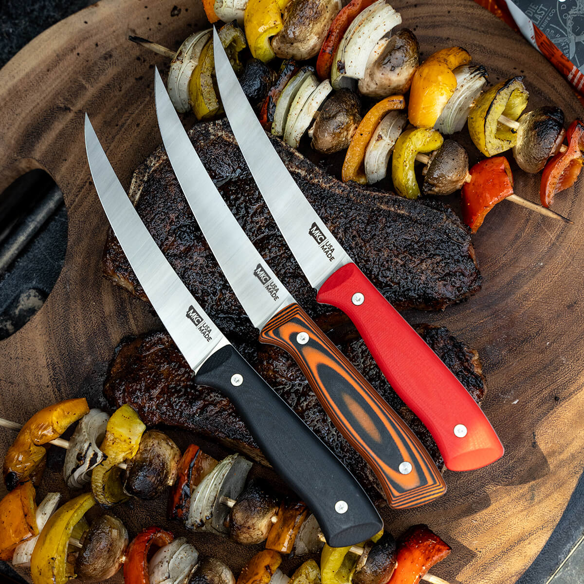 Alt text: "Three Bearded Butchers boning butcher knives with sharp, pointed blades and ergonomic handles in black, red, and orange displayed on a wooden board with barbecued steak and grilled vegetables."