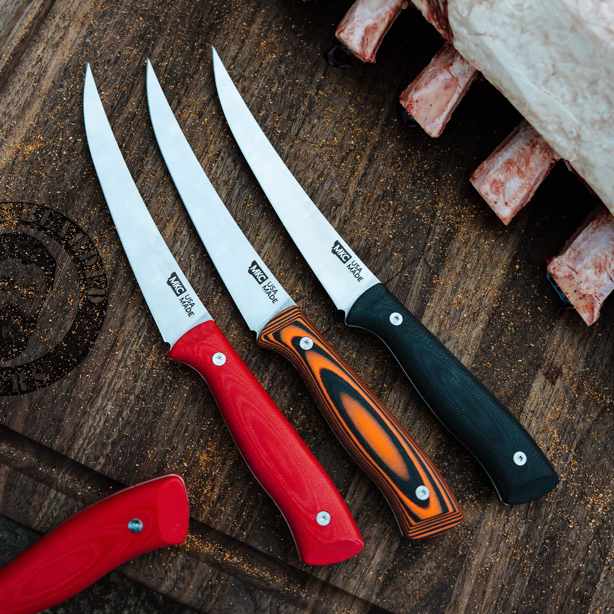 Three Bearded Butchers boning butcher knives with sharp, pointed blades, featuring different handle colors including red, black, and striped orange and black, displayed on a rustic wooden surface with meat cuts in the background.