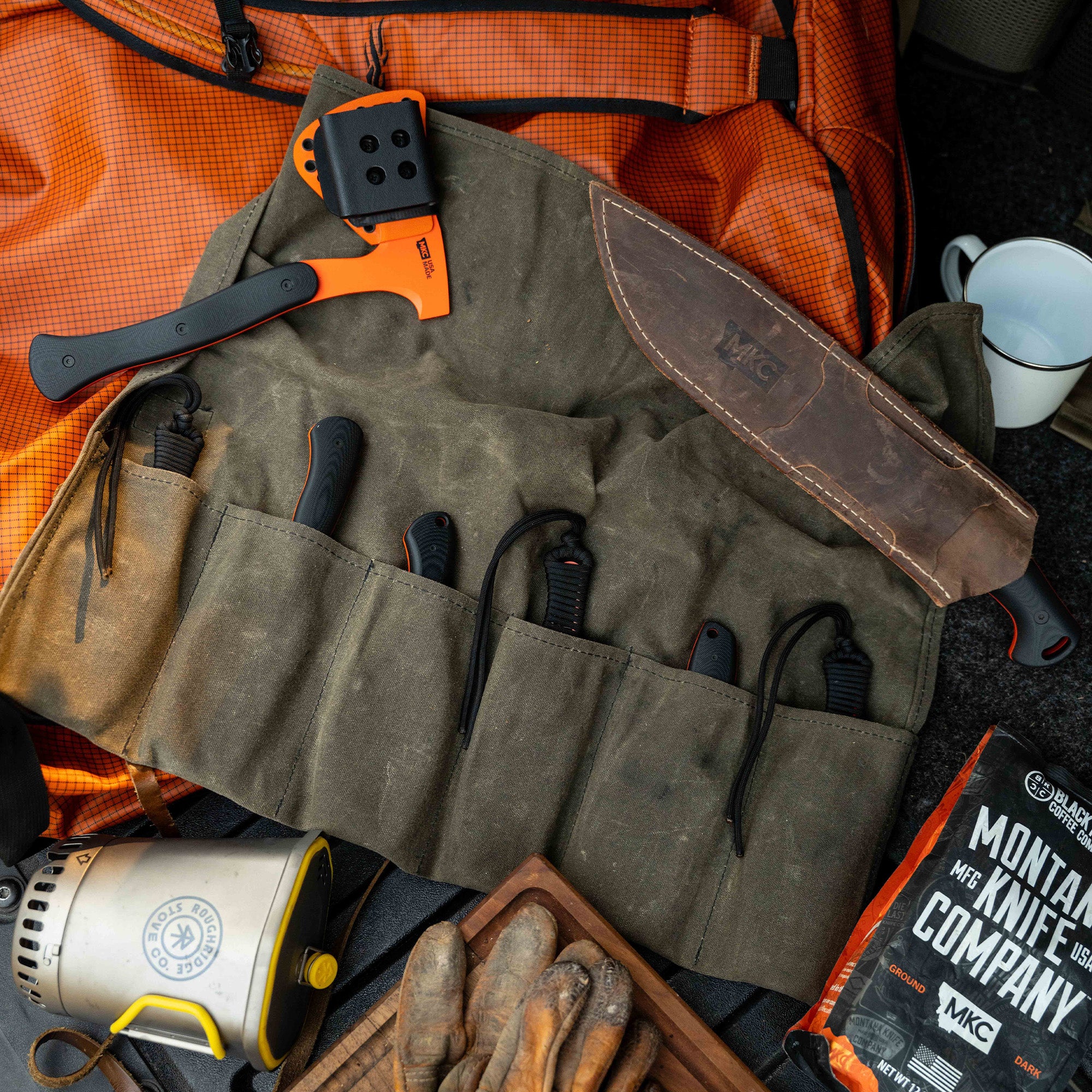Full set of eight Blaze Orange Cerakote knives displayed on a canvas roll, alongside outdoor gear and tools.