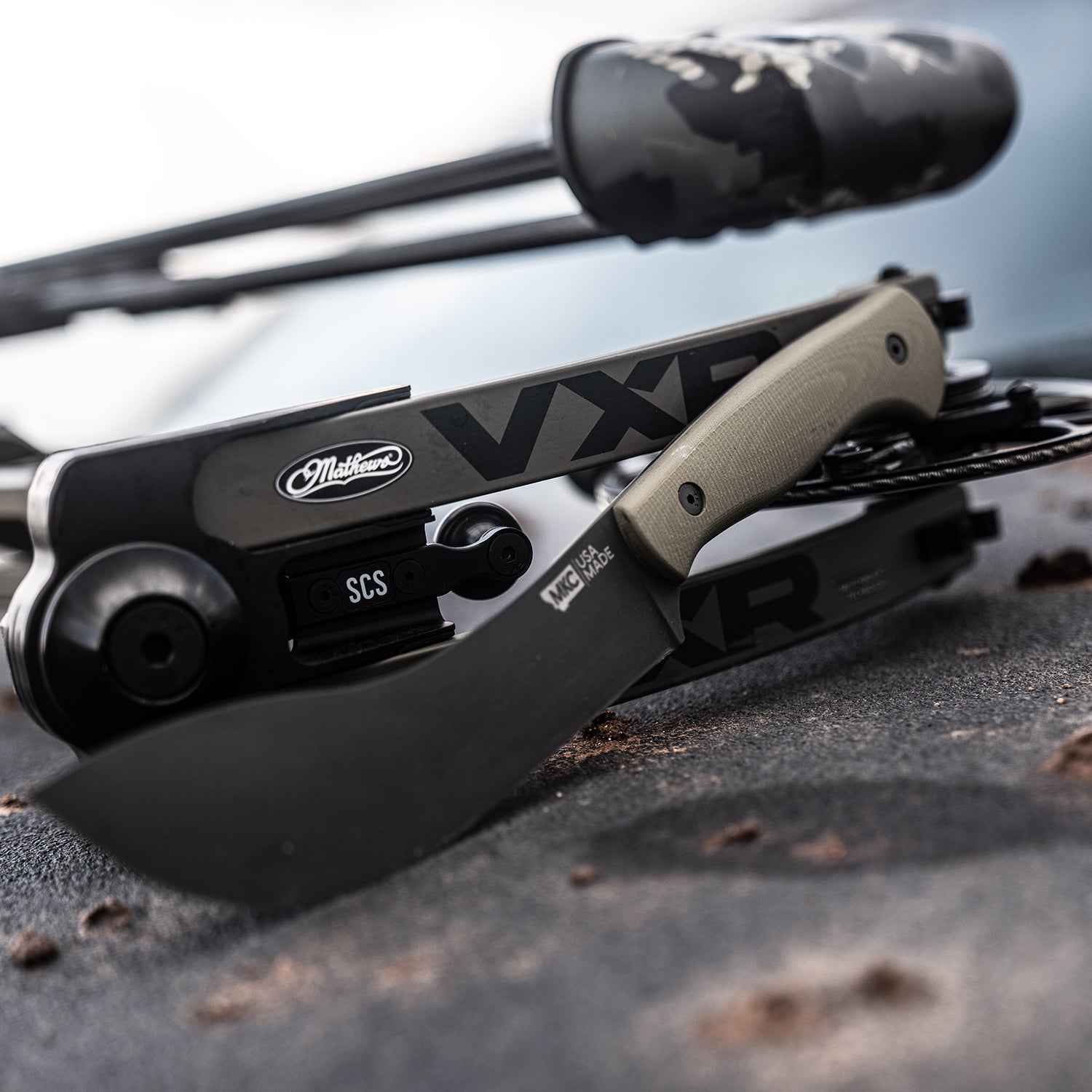 Alt text: Close-up view of a Beartooth Pro Skinning Blade with an olive handle, prominently displayed against a blurred background of outdoor equipment. The blade is curved with a matte black finish, featuring visible branding and mounted on a gear setup.