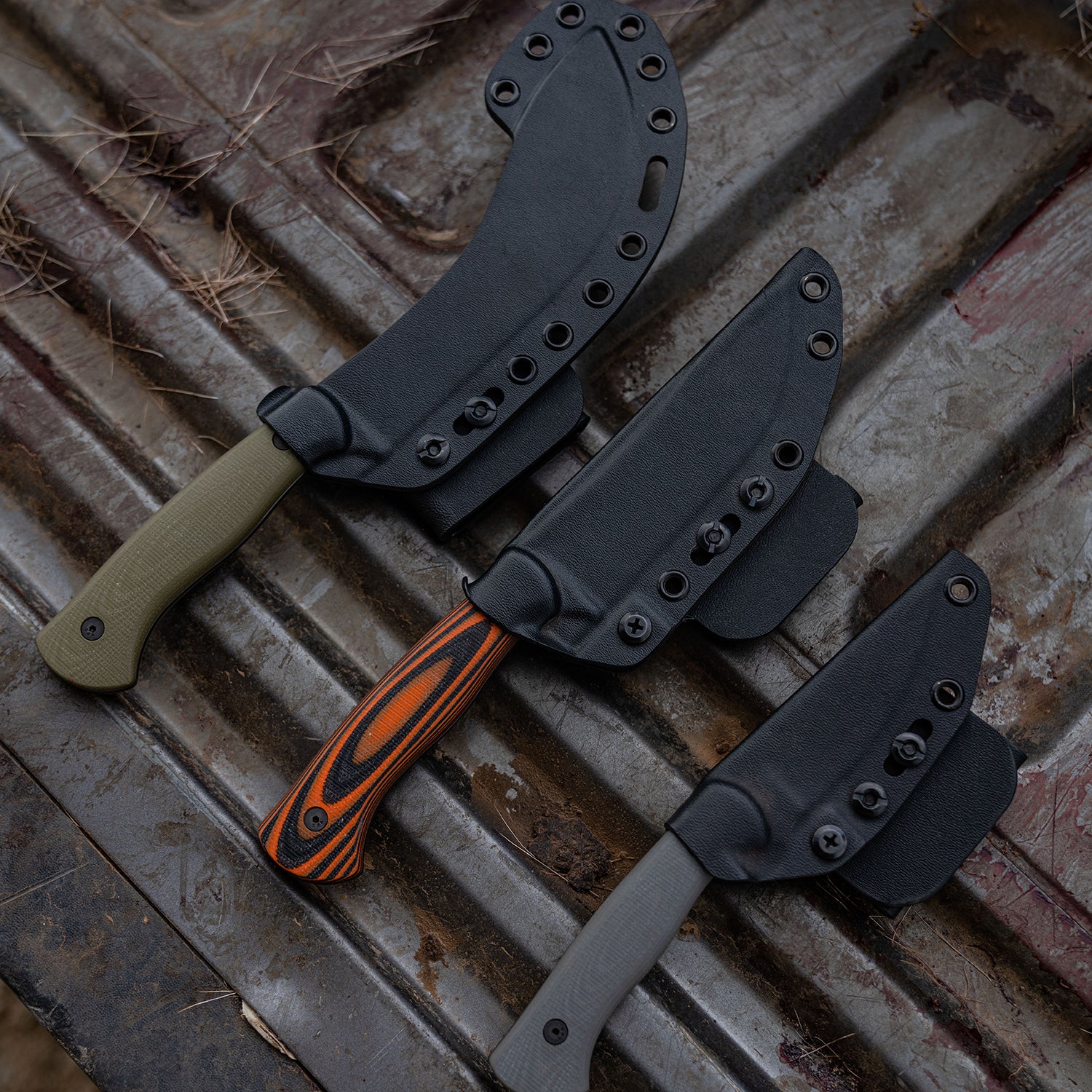 Alt text: "A set of Beartooth Pro Skinning Blades with olive and orange handle options, each knife sheathed in a black holster, displayed on a rustic metallic surface."