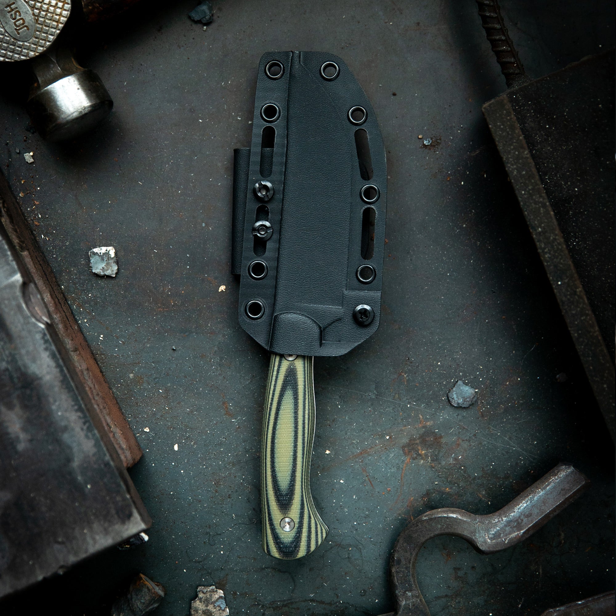 Alt text: "Magnacut Stonewall Skinner knife with a detailed green and black striped handle, partially sheathed in a black textured holster, displayed on a dark, gritty metal surface alongside industrial tools."