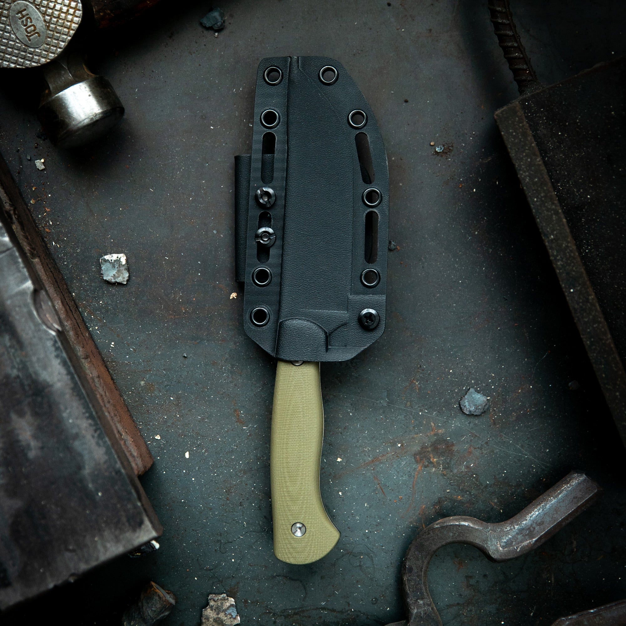 Alt text: Close-up of a MagnaCut Stonewall Skinner knife by Mule Deer Foundation, featuring an olive green handle and a black sheath, displayed on a grungy metal surface alongside vintage tools.