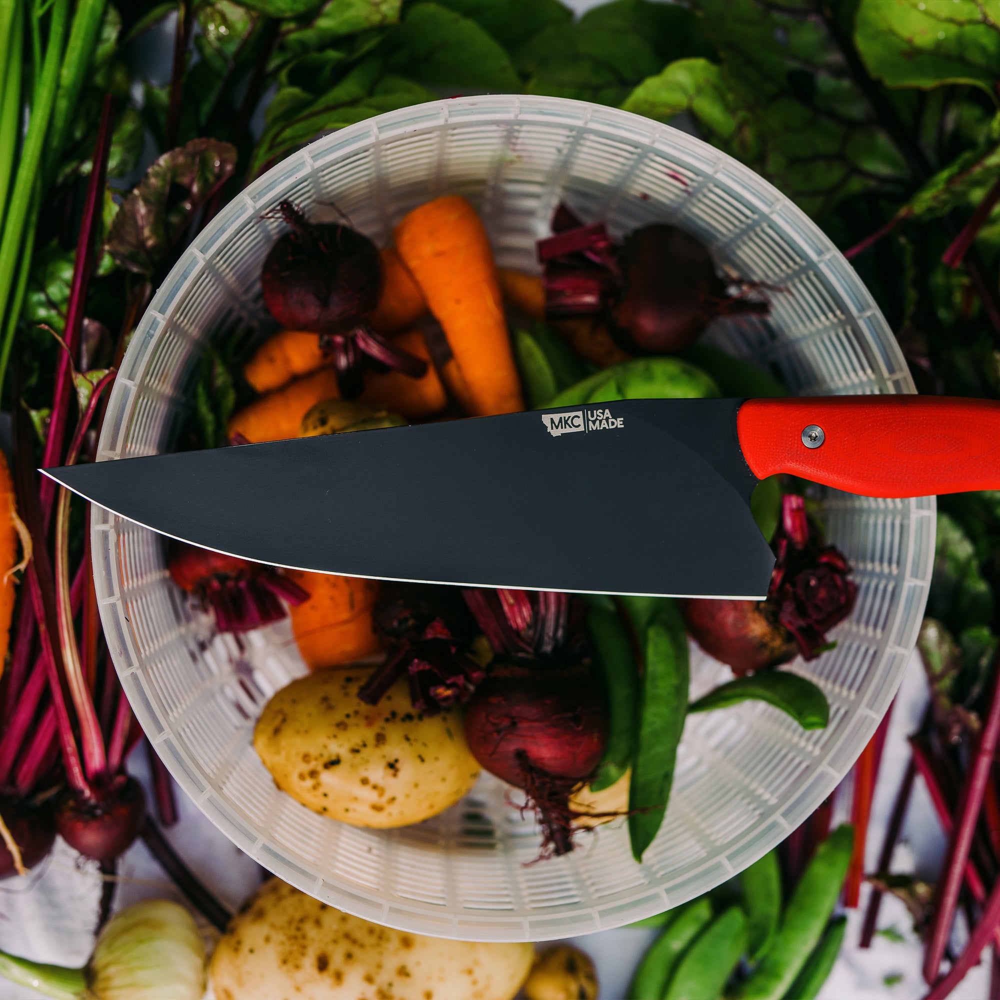 Alt text: "A BIGHORN CHEF Red Chef Knife with a sleek black blade and vibrant red handle, displayed on a clear bowl filled with an assortment of fresh vegetables including carrots, beets, and leafy greens."