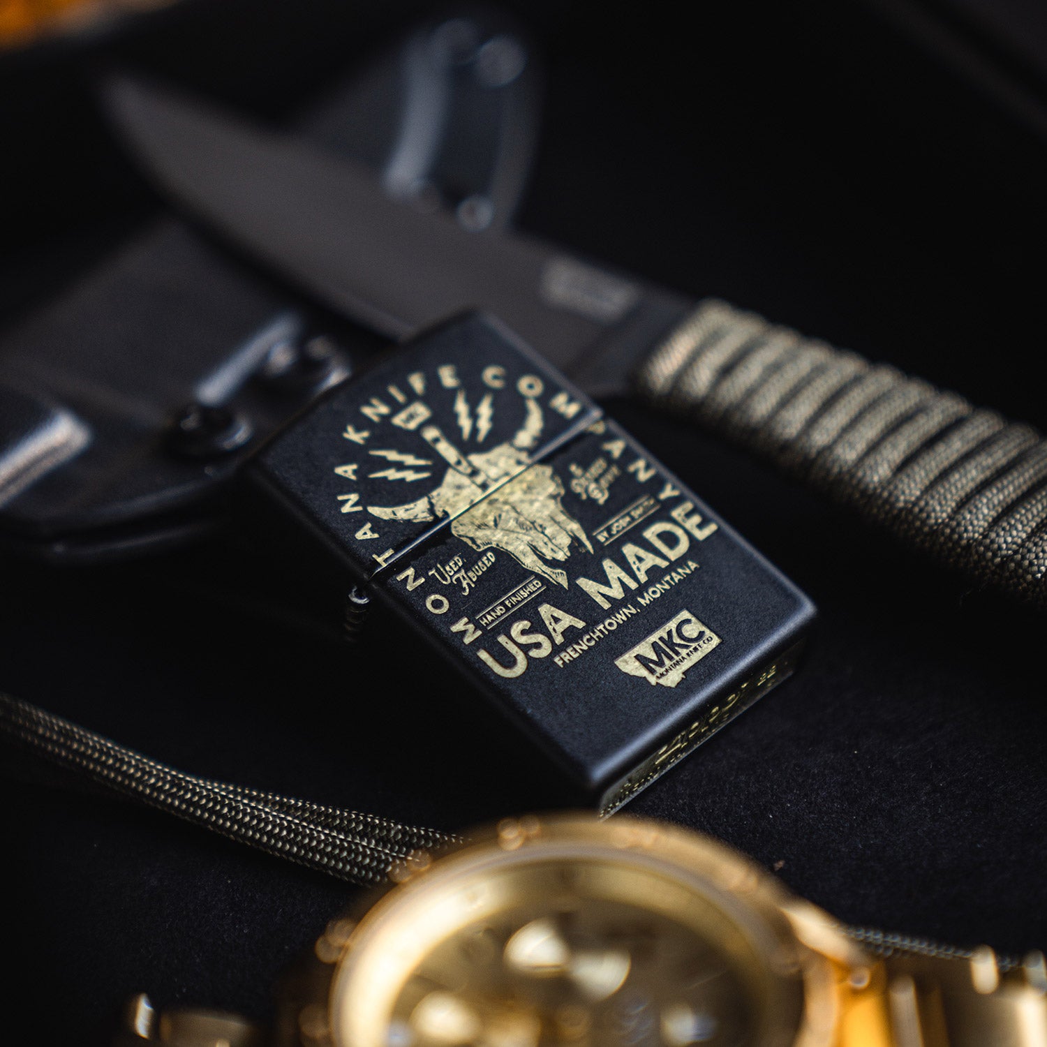 Alt text: "Close-up of a black windproof Zippo lighter featuring a bison skull design and text 'USA MADE'. The lighter is displayed on a dark background alongside a knife and a gold watch."