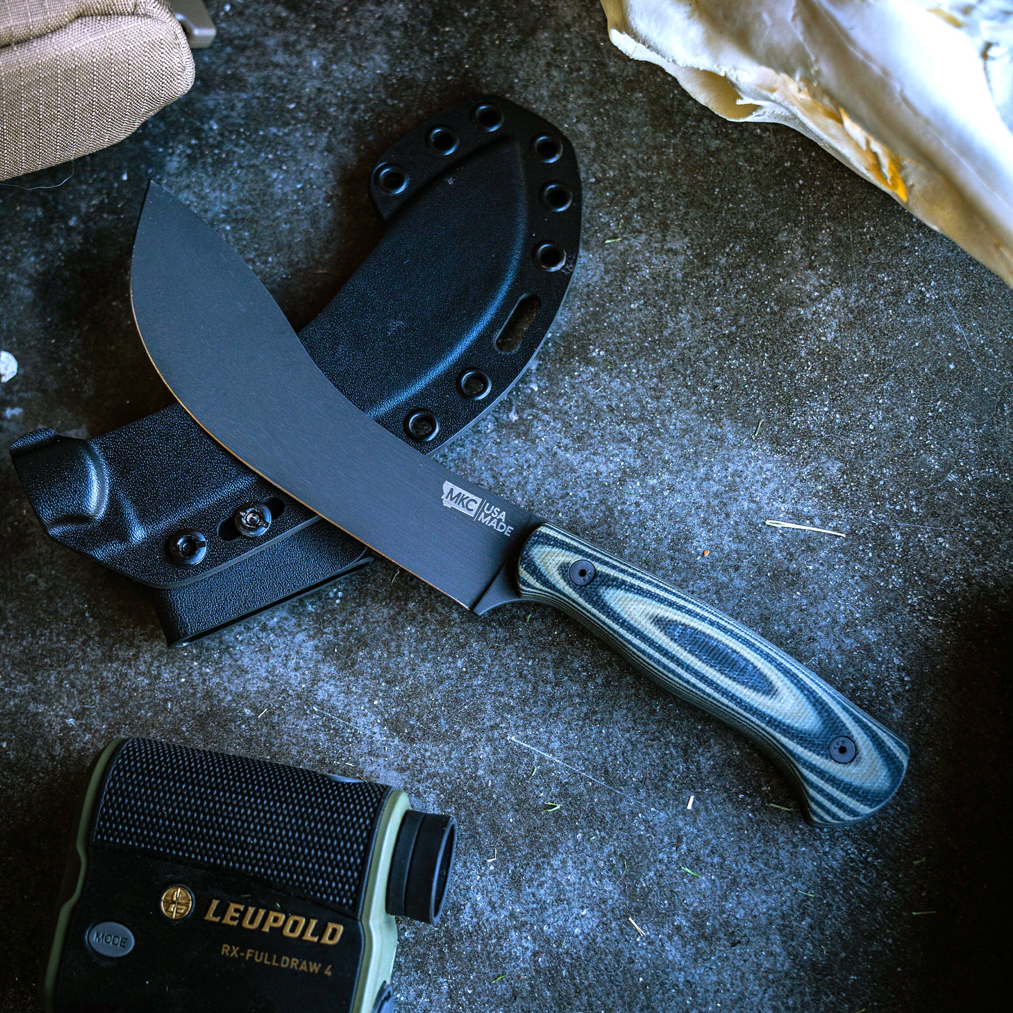 Alt text: A Beartooth Pro Skinning knife with a black blade and green and black striped handle, displayed next to its black sheath on a textured concrete surface, with outdoor gear in the background.