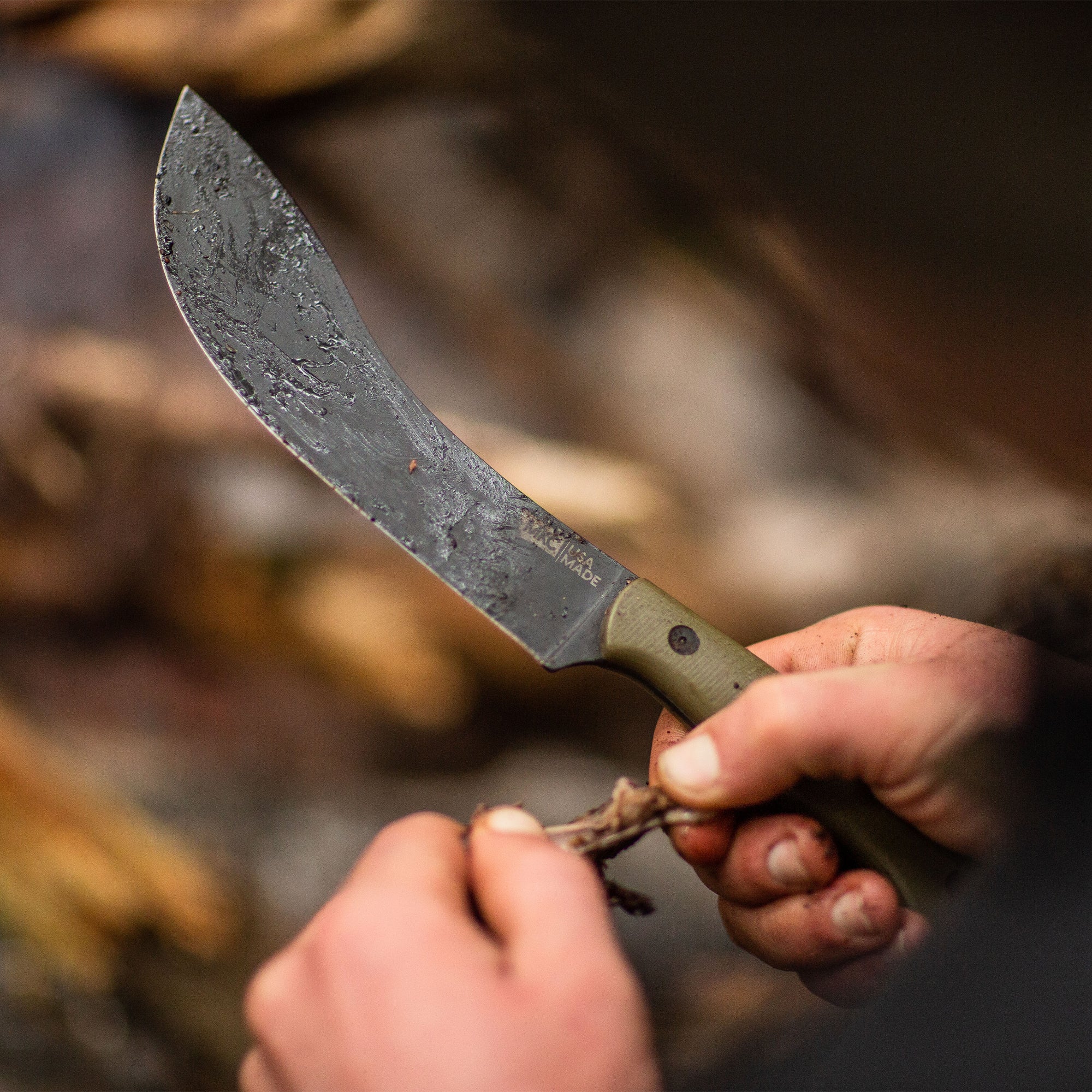Close-up image of a person holding a Beartooth Pro Skinning Blade with an olive-colored handle, demonstrating its use on a piece of wood in a natural setting.