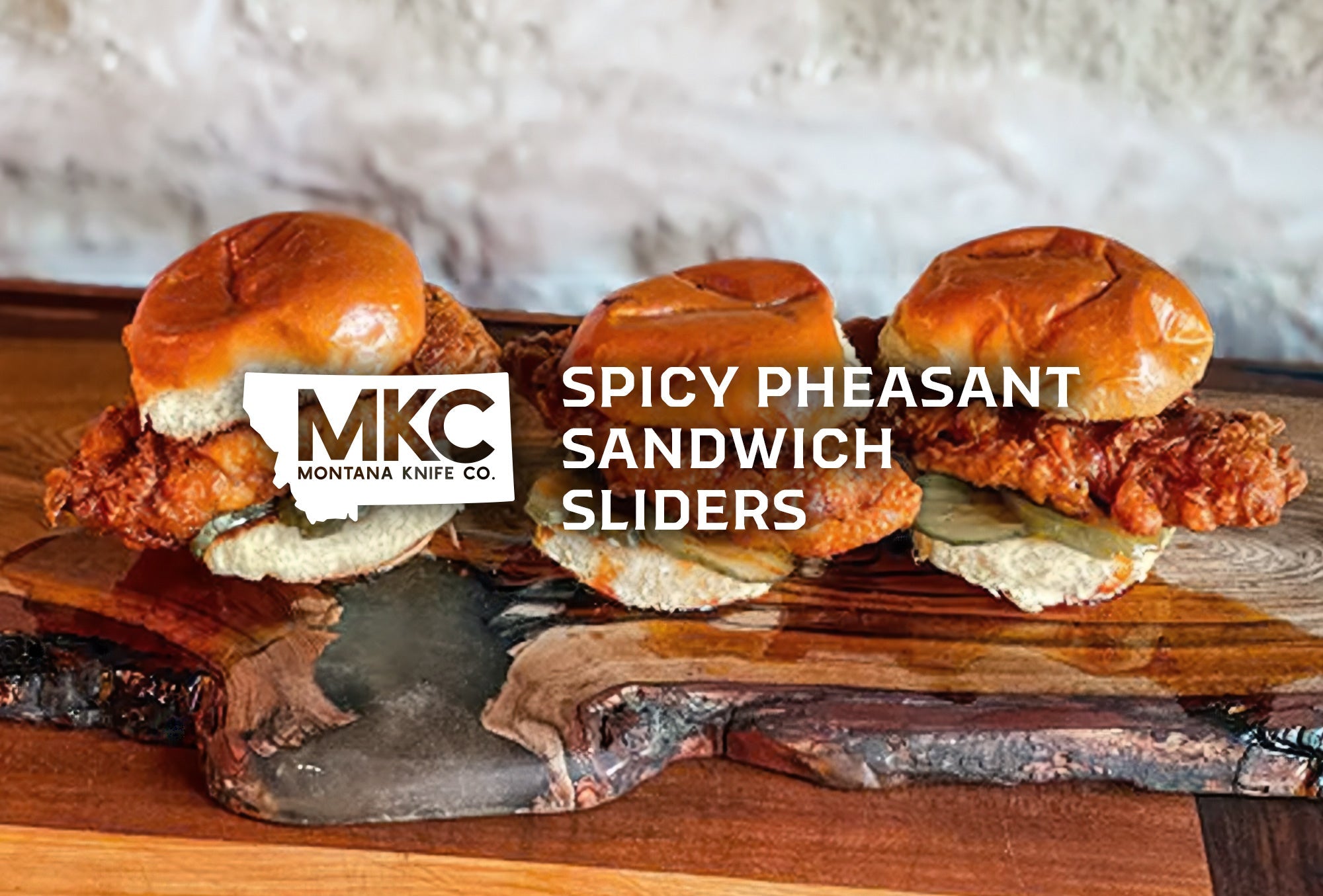 Three golden, crispy, spicy pheasant sandwich sliders topped with pickles resting on a wooden surface.