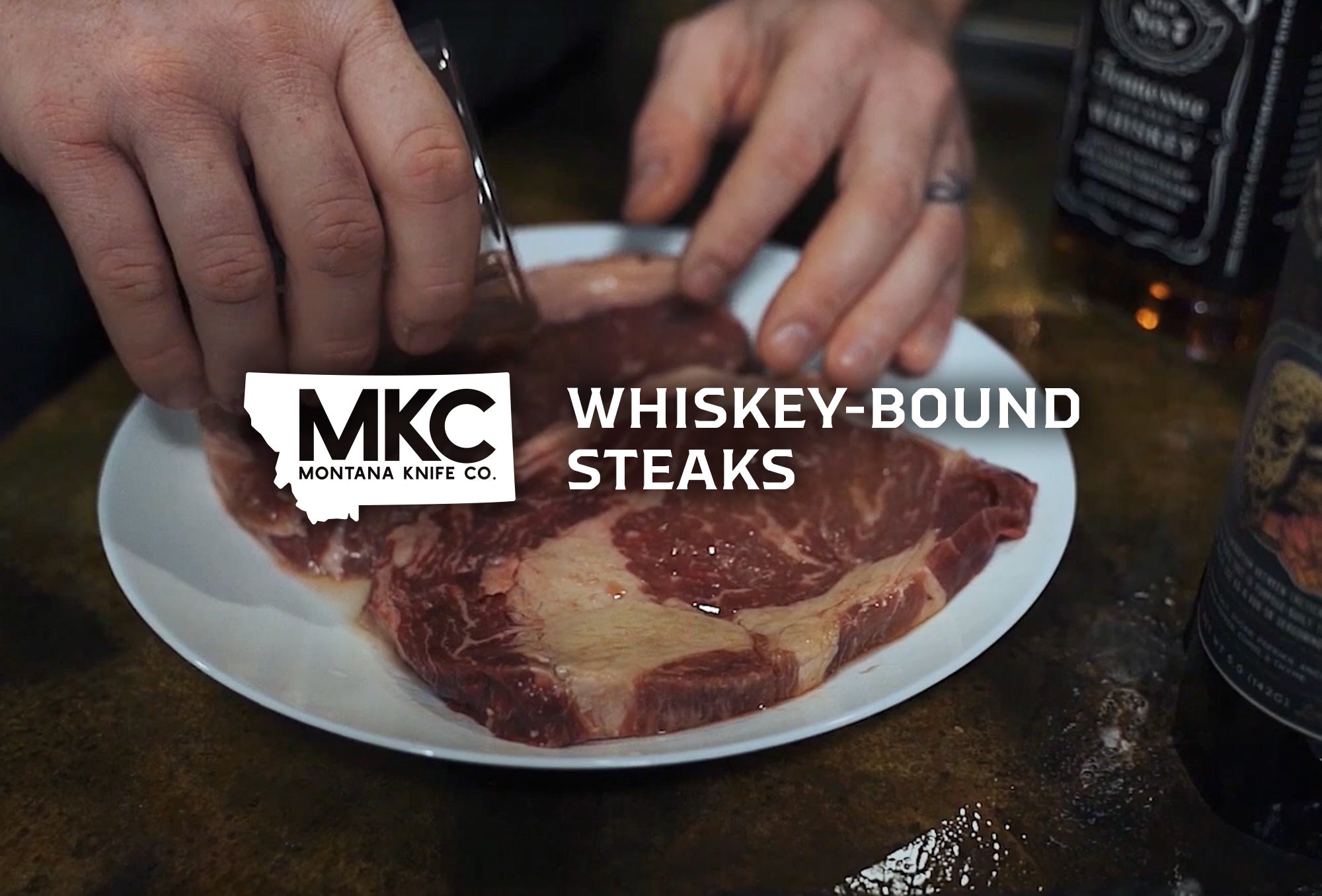 Hands using a knife to prepare raw ribeye steaks on a white plate with whiskey bottle in the background.