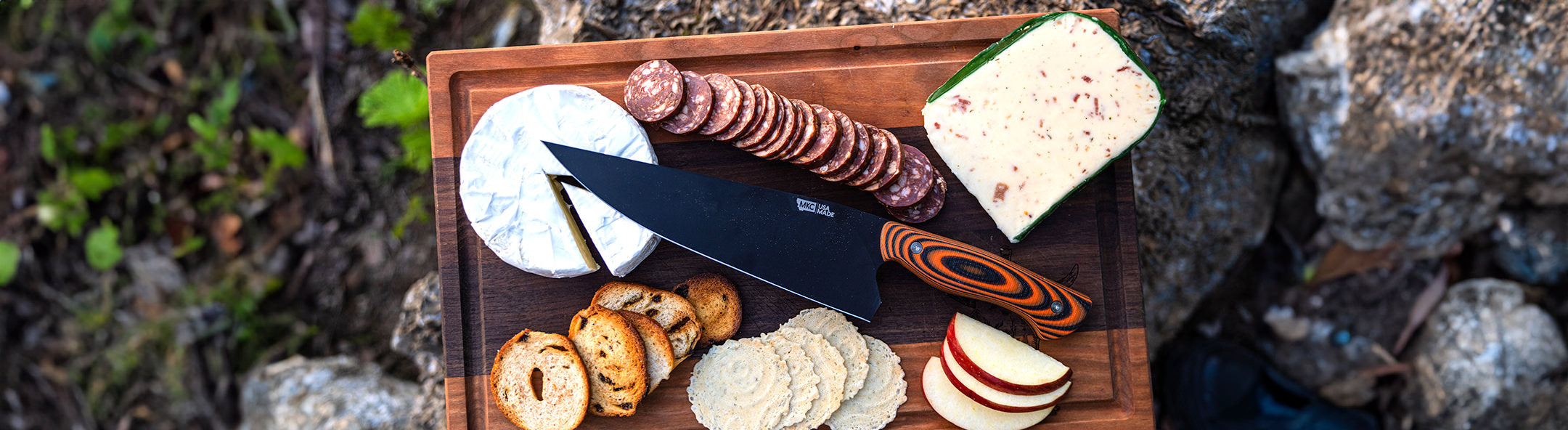 Chef knife with meats and cheeses on wooden cutting board.