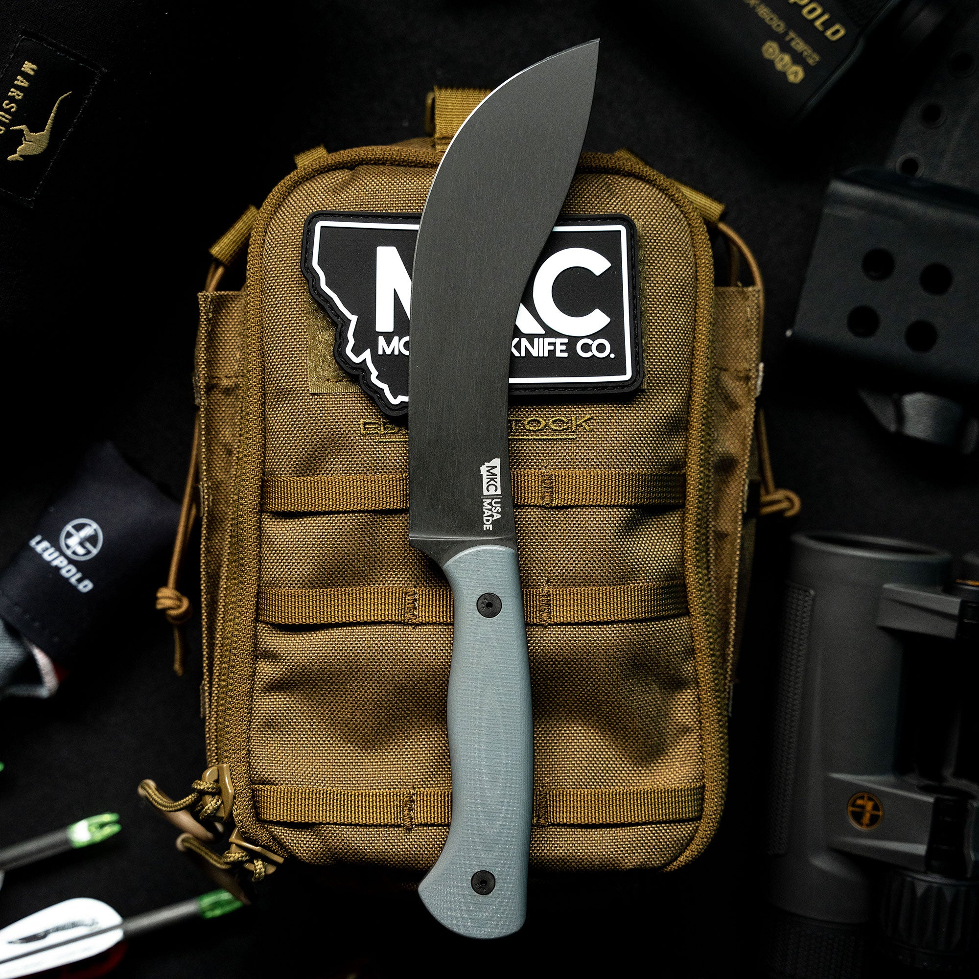 A Beartooth Pro Skinning Blade in grey with a lightweight handle, displayed vertically against a brown tactical backpack, surrounded by outdoor gear.
