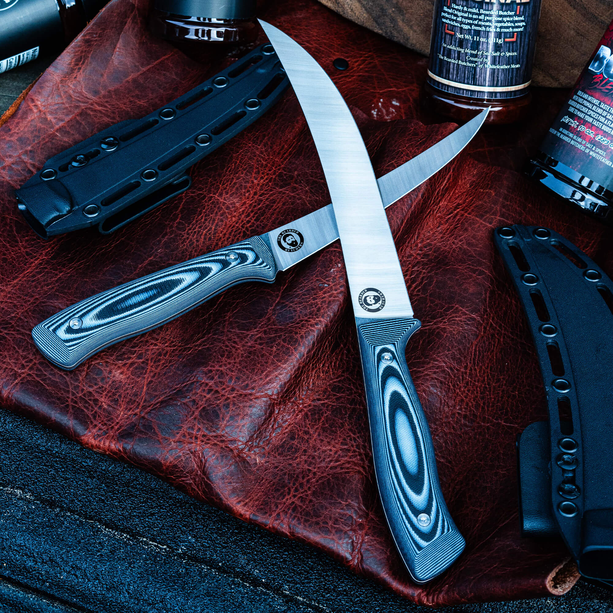 Alt text: High-quality breaking butcher knives with intricately patterned handles displayed on a textured red leather surface, accompanied by black sheaths and outdoor cooking products. The blades feature reflective silver surfaces and branded logos.