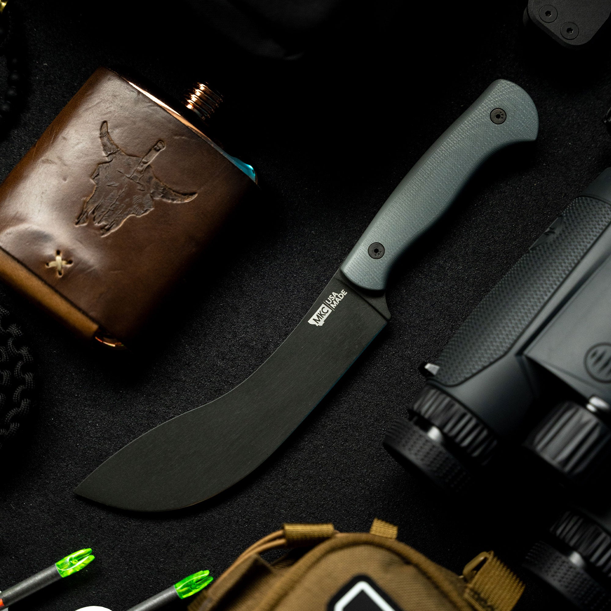 Alt text: "Beartooth Pro Skinning Blade in grey, featuring a curved blade and a textured handle, displayed alongside tactical gear including a flint, a brown leather flask, and a backpack on a dark background."