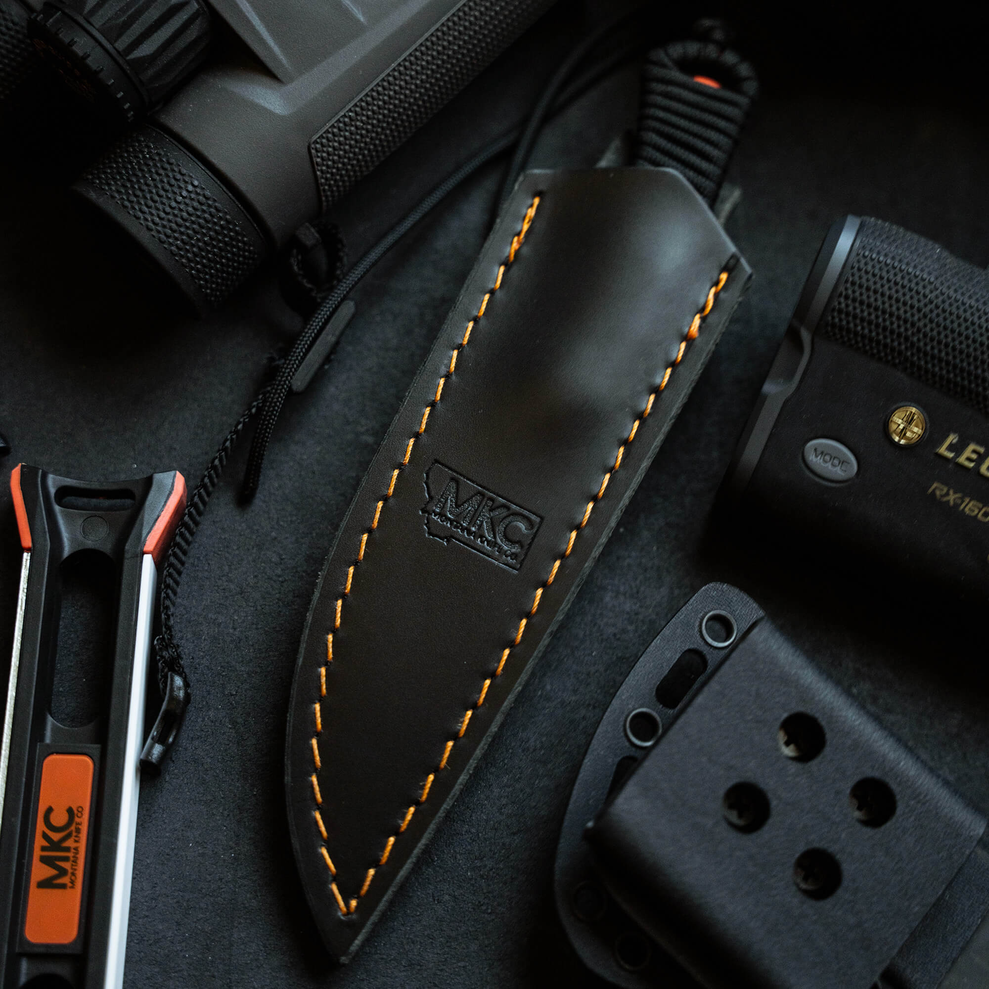 Black leather sheath with orange stitching for vertical belt carry, prominently displaying the MKC logo, surrounded by outdoor gear including a flashlight and multitool.