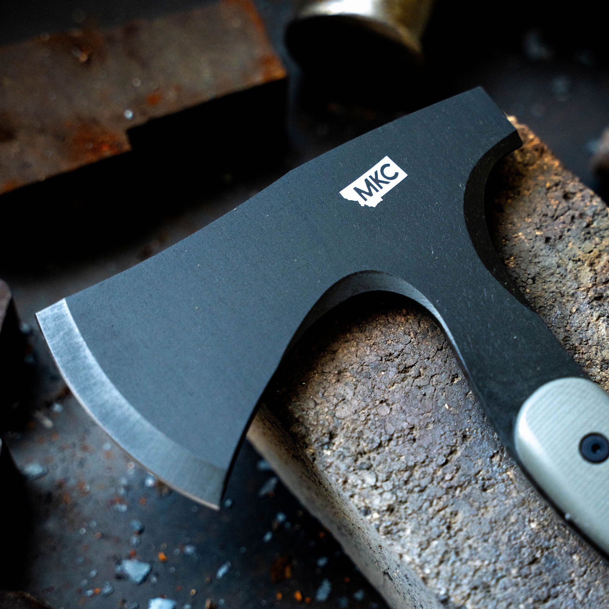 Close-up of a MKC AEV Hellgate Hatchet with a black matte finish and sharp blade, resting on a worn industrial surface