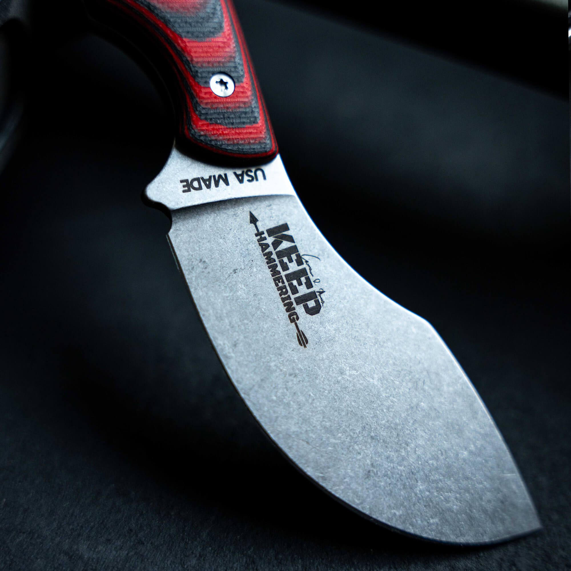 Close-up of a Keep Hammering Packout Skinner knife with a red and black handle and a stonewashed blade, featuring the logo and text detail.