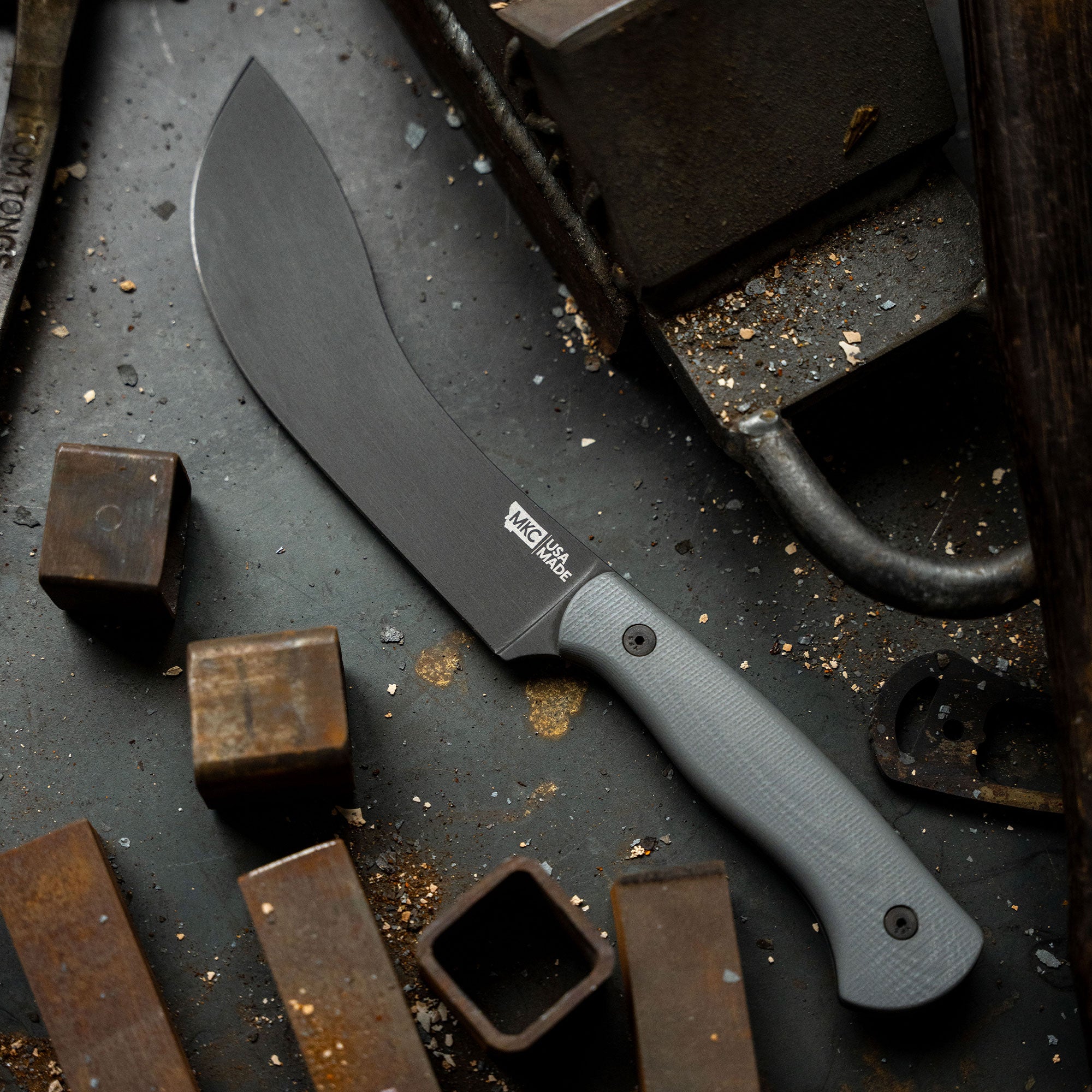 Alt text: "Beartooth Pro Skinning Blade in grey with a curved matte black blade and textured grey handle, displayed on a dark, gritty surface surrounded by metal objects and debris."