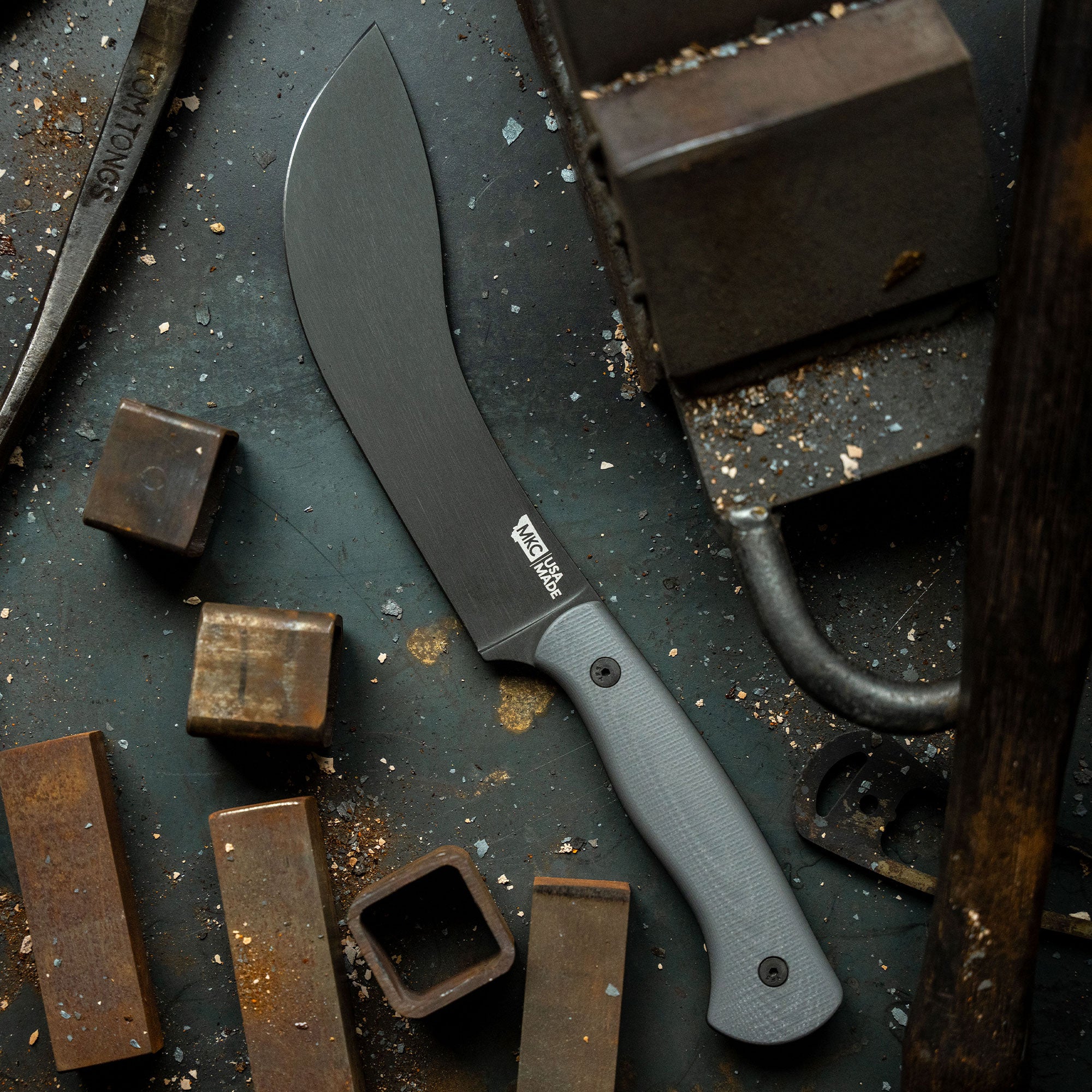 Alt text: Grey Beartooth professional skinning knife with a matte finish and a textured handle, displayed on a grungy workbench surrounded by metal blocks and tools, highlighting the blade's robust construction for outdoor activities.