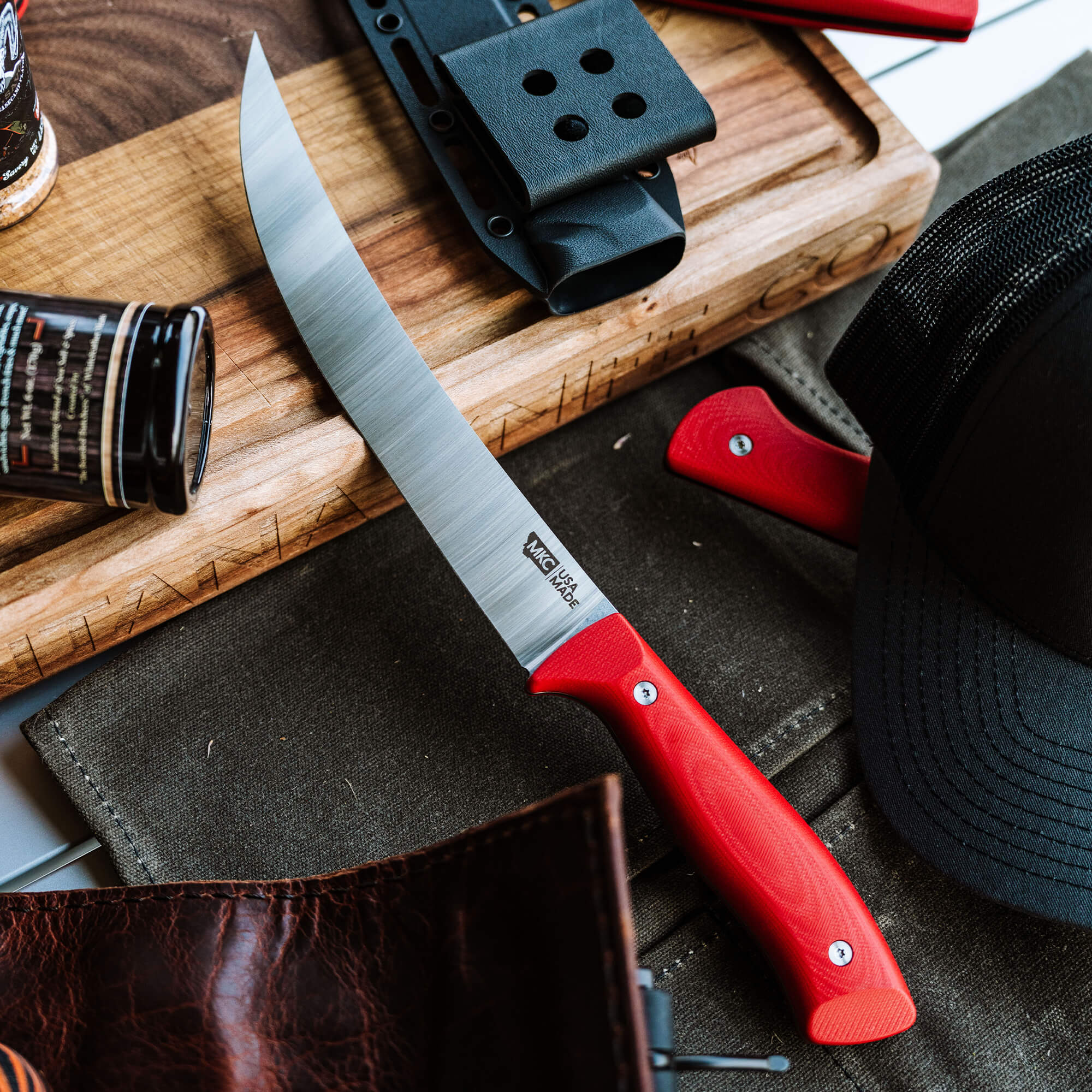 Alt text: Long chef's knife with a red handle and a matte black sheath, displayed on a wooden cutting board alongside accessories like a black cap and a brown leather wallet, embodying a rugged culinary theme.