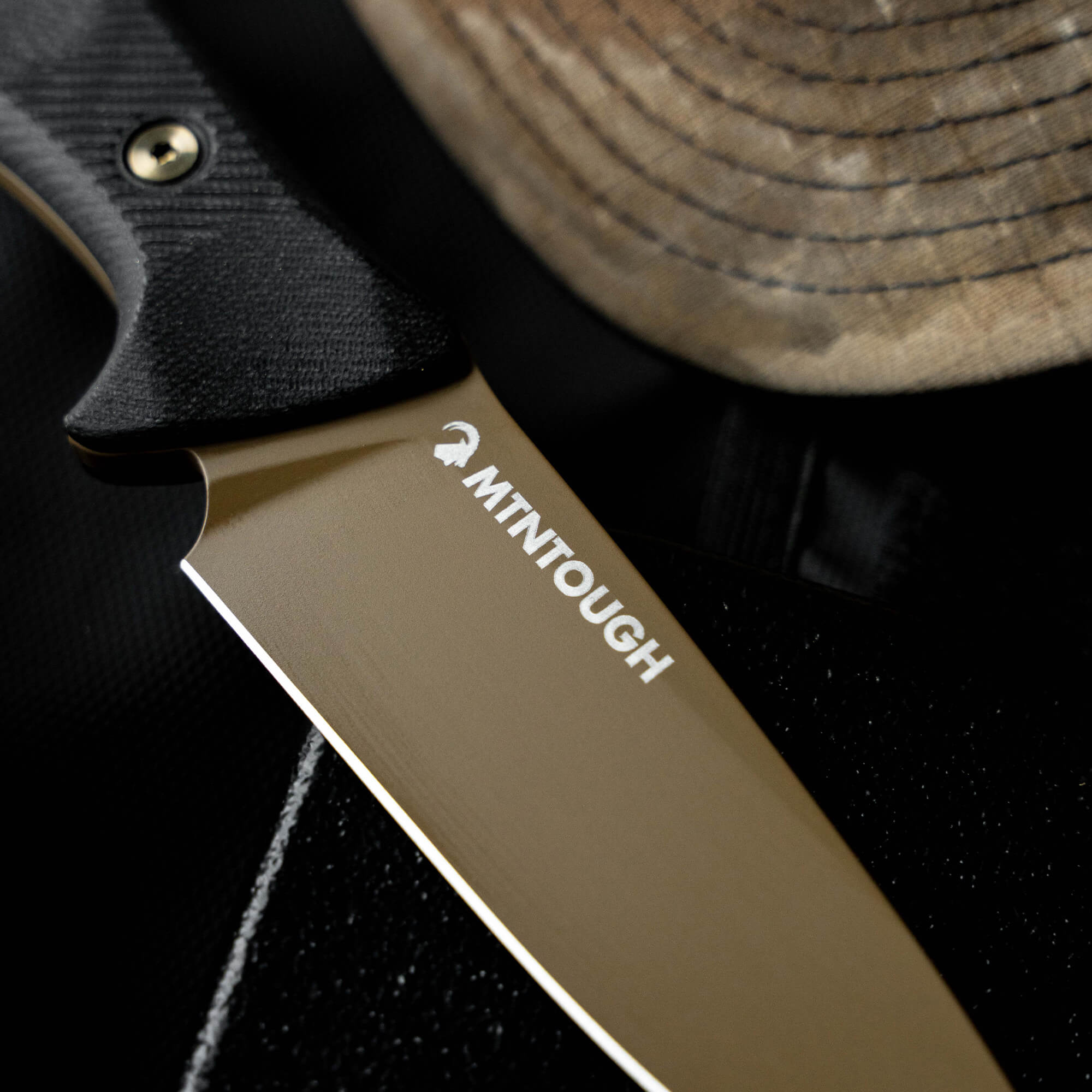 MTNTOUGH TF24 knife with a sleek blade and textured handle, resting beside a worn hat on a dark surface.
