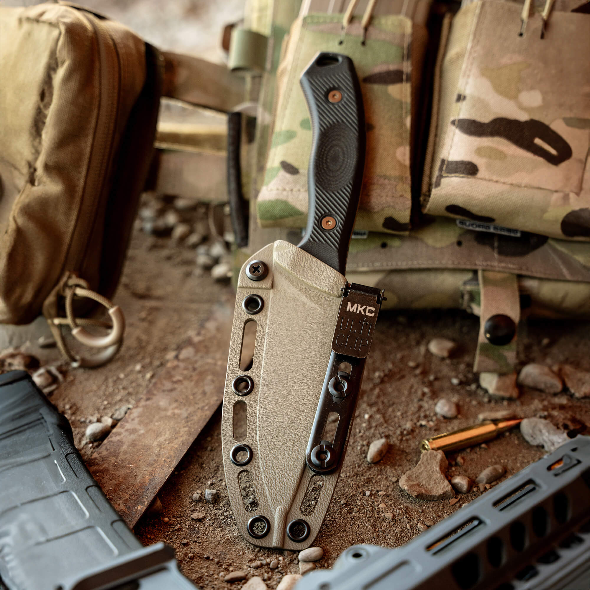 Alt text: "Close-up of a coyote tan Kydex sheath with a tactical knife secured inside, prominently displayed against a backdrop of military gear and rugged outdoor terrain."