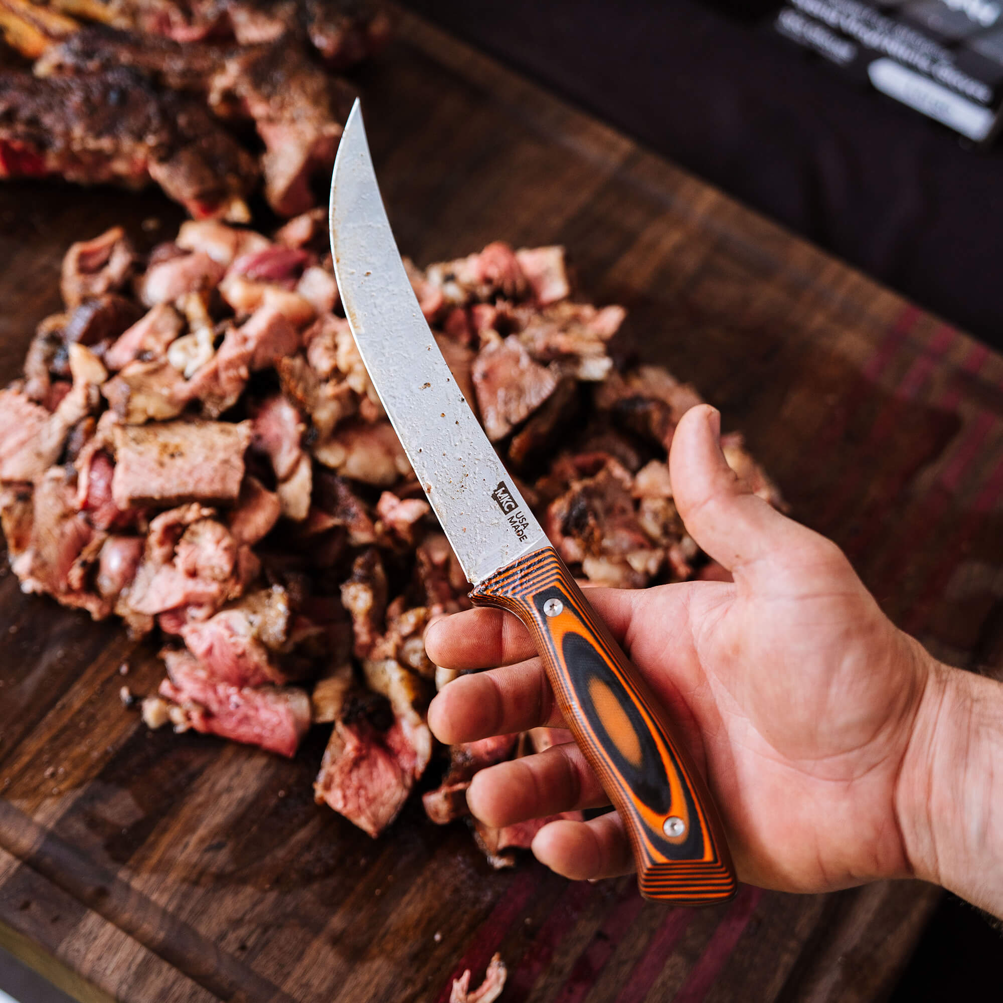 Alt text: "A hand holding a Bearded Butchers breaking butcher knife with an orange and black handle, over a wooden board covered in chopped meat pieces."