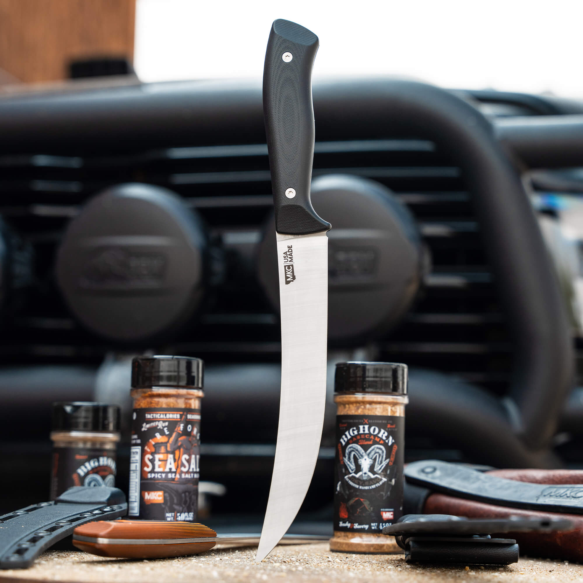 Long, narrow Bearded Butchers breaking butcher knife with a black handle, displayed upright in front of a vehicle, surrounded by various seasoning containers and butchery tools.