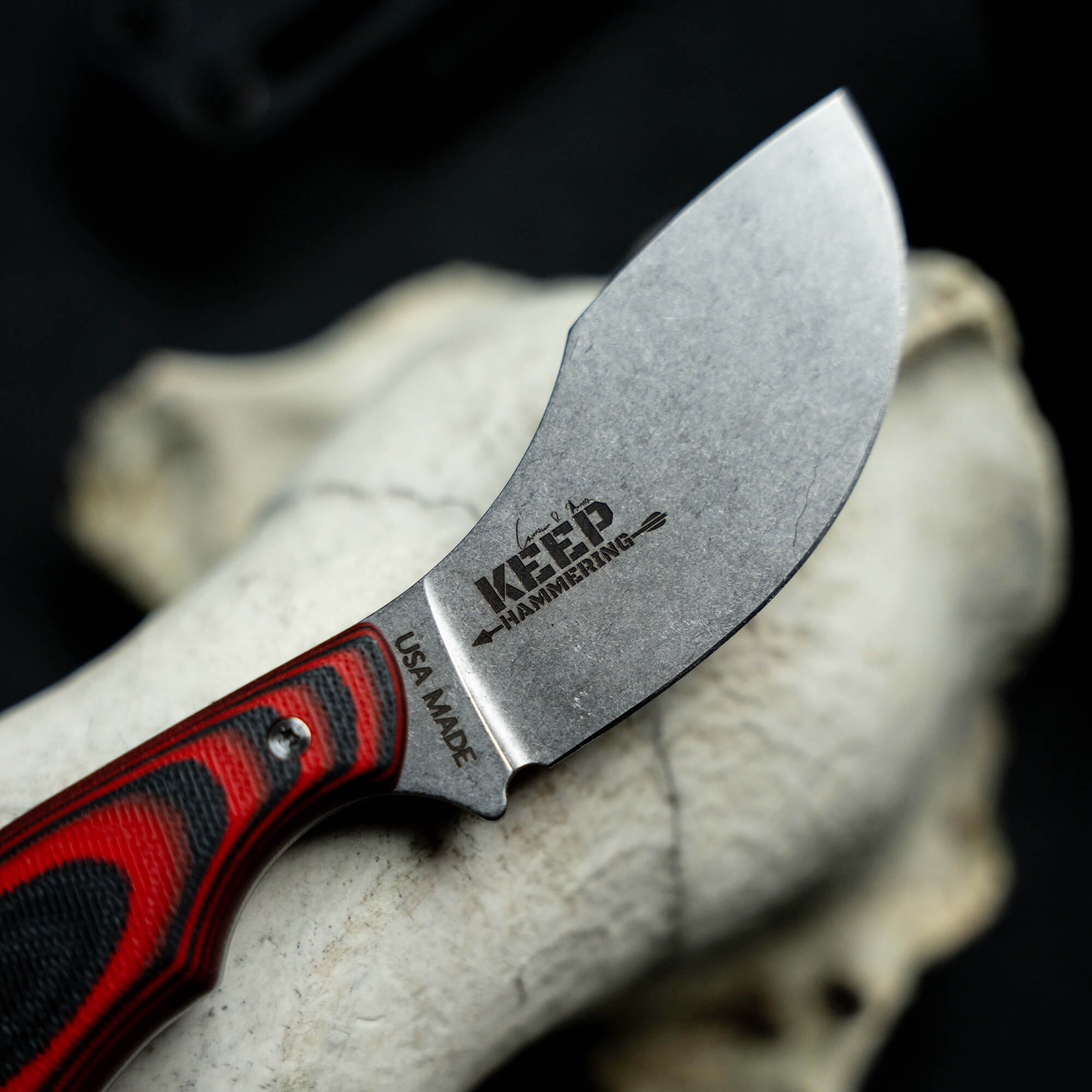 Close-up of a 'Keep Hammering Packout Skinner' knife with a red and black handle, showcasing its blade marked with "KEEP HAMMERING" and "USA MADE". The knife is displayed against a black background with a bone element visible
