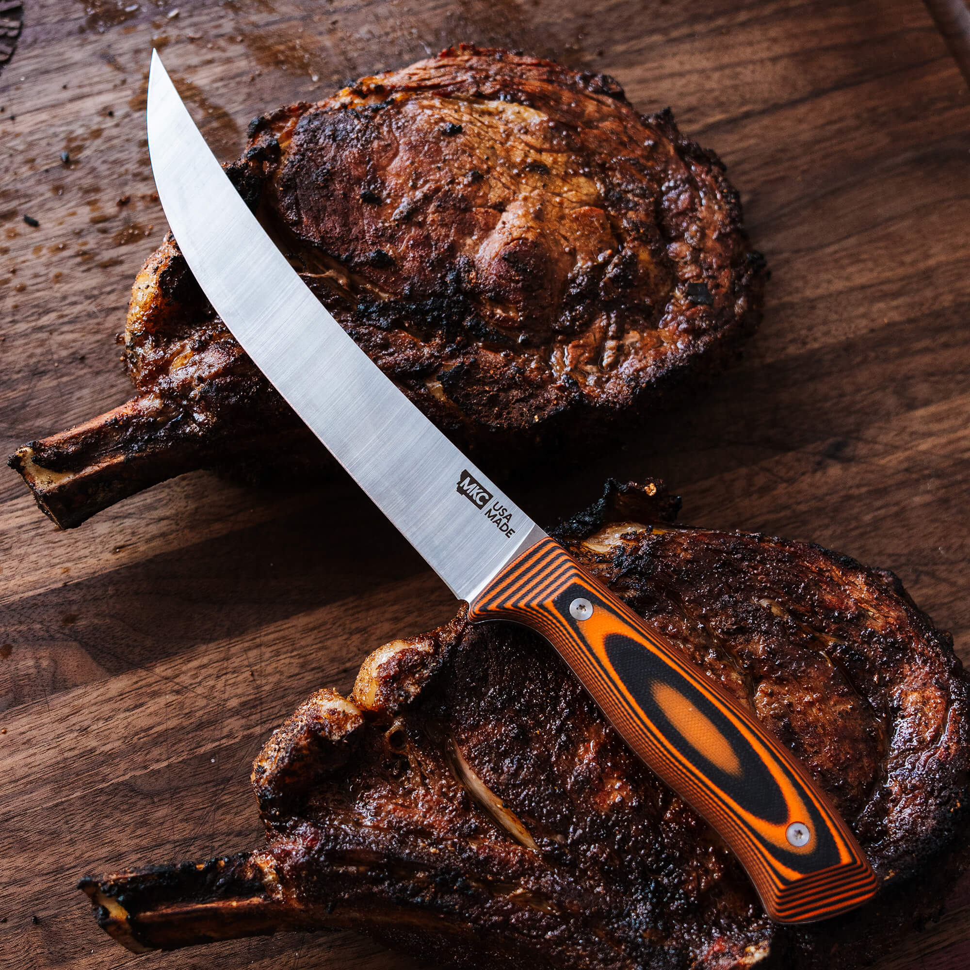 Descriptive alt text: "Breaking Butcher knife by Bearded Butchers with a laminated orange and black handle, prominently displayed on a succulent, well-seared steak on a wooden table."