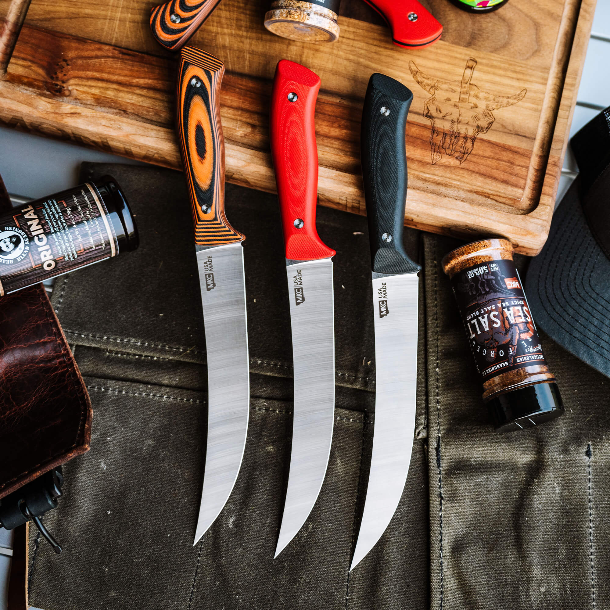 Three Bearded Butchers brand boning knives with sleek designs, featuring orange, red, and black handles, displayed on a wooden cutting board with seasoning containers nearby.