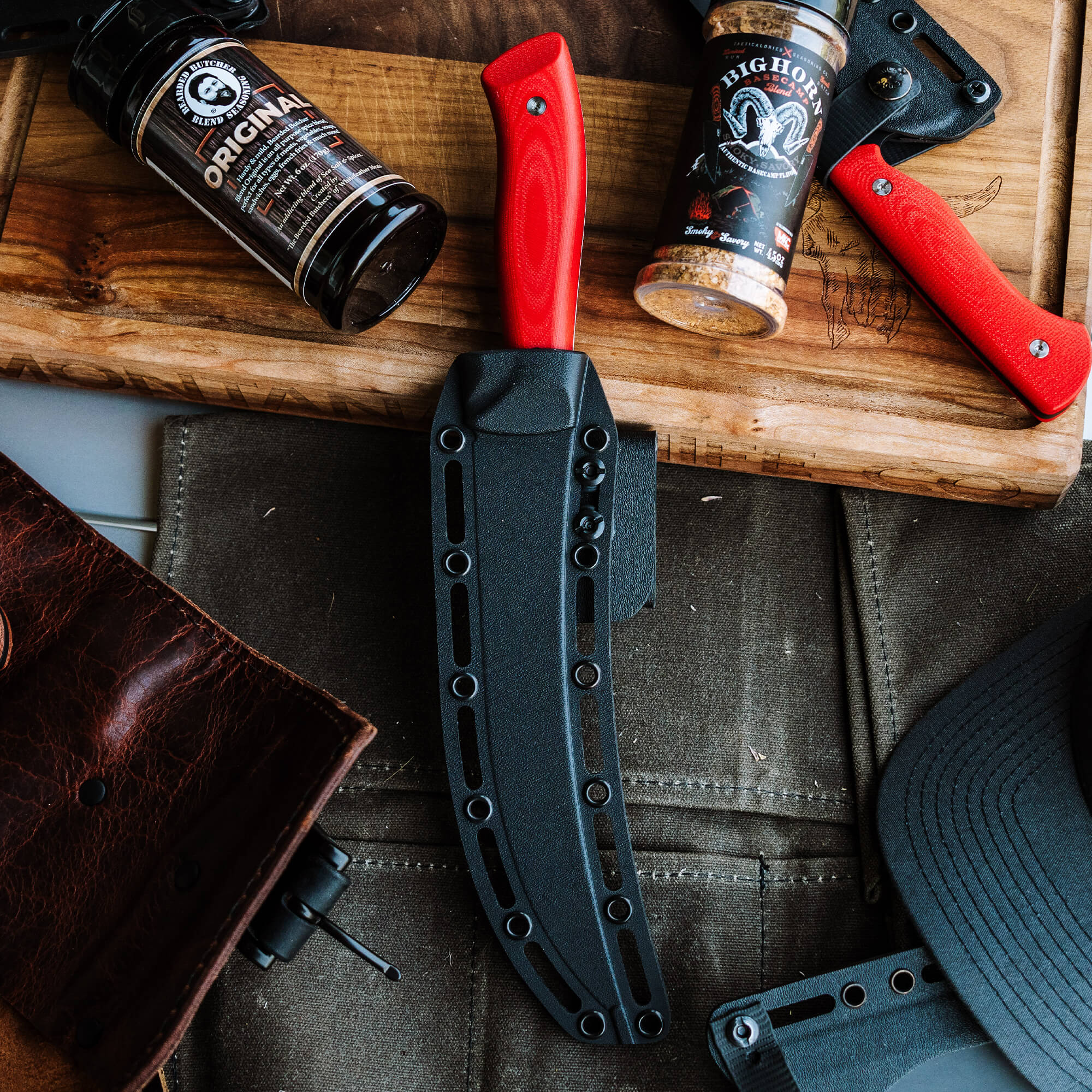 Breaking Butcher knife with red handle and black sheath, displayed on a wooden board alongside a leather wallet, seasoning bottles, and cap, indicating a lifestyle theme for Bearded Butchers products.