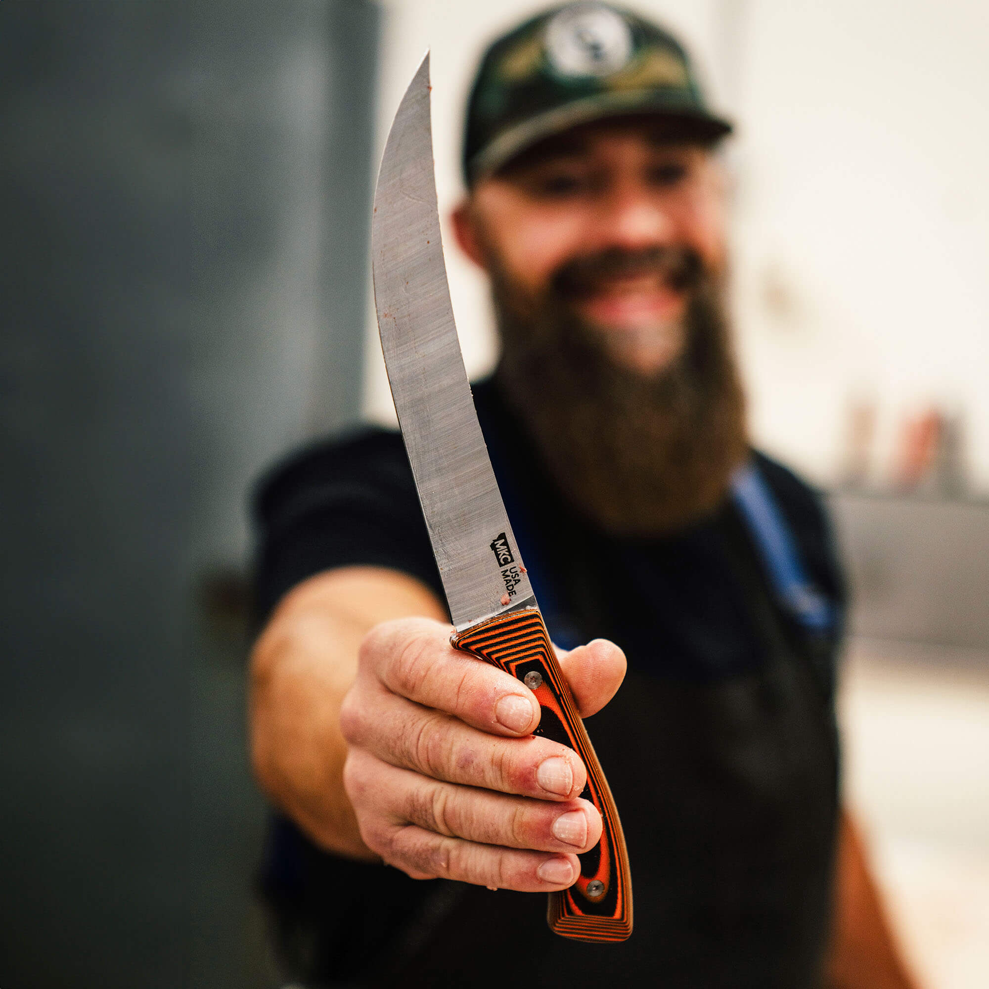 Smiling bearded man in a cap and apron presenting a large butcher knife with an orange handle, implying quality and precision in meat processing.