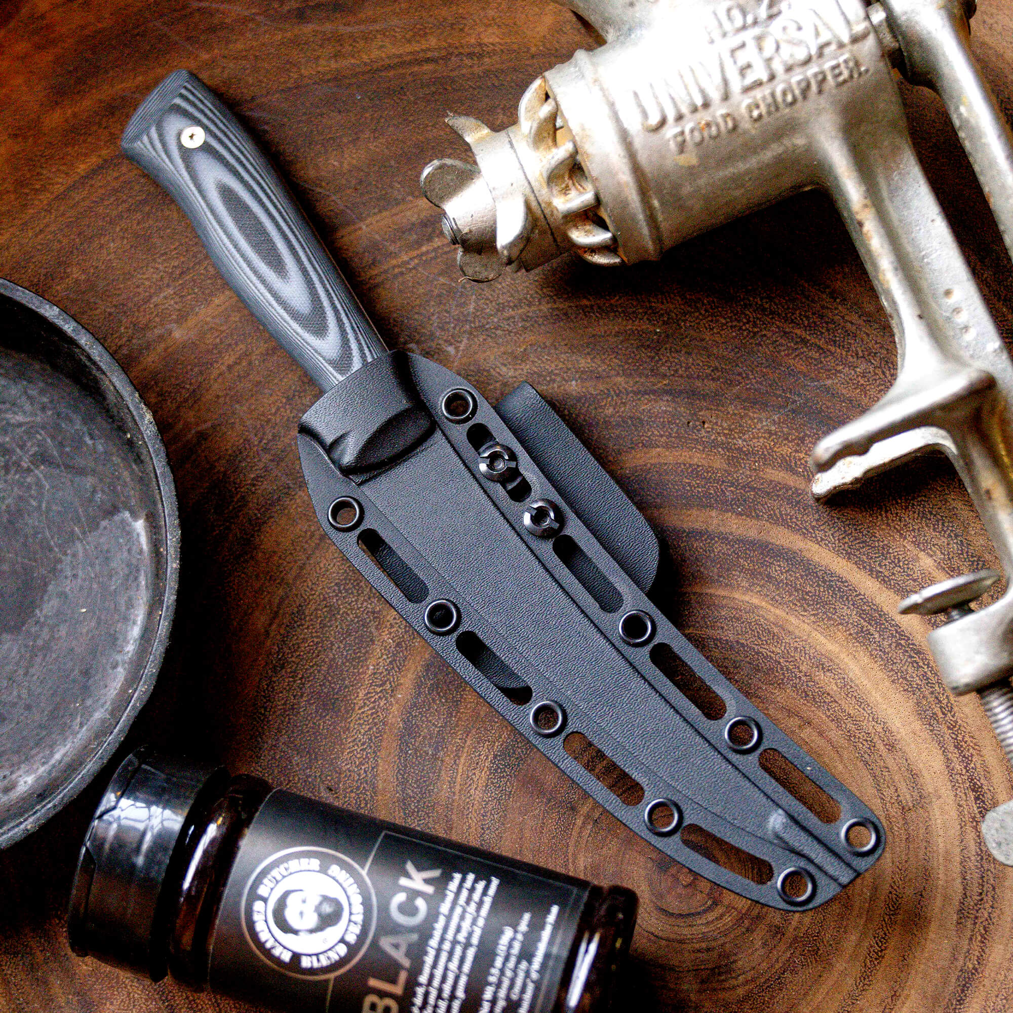 Alt text: A Bearded Butchers boning butcher knife with a black handle and sheath displayed on a wooden surface, alongside an antique food chopper and a bottle of seasoning.