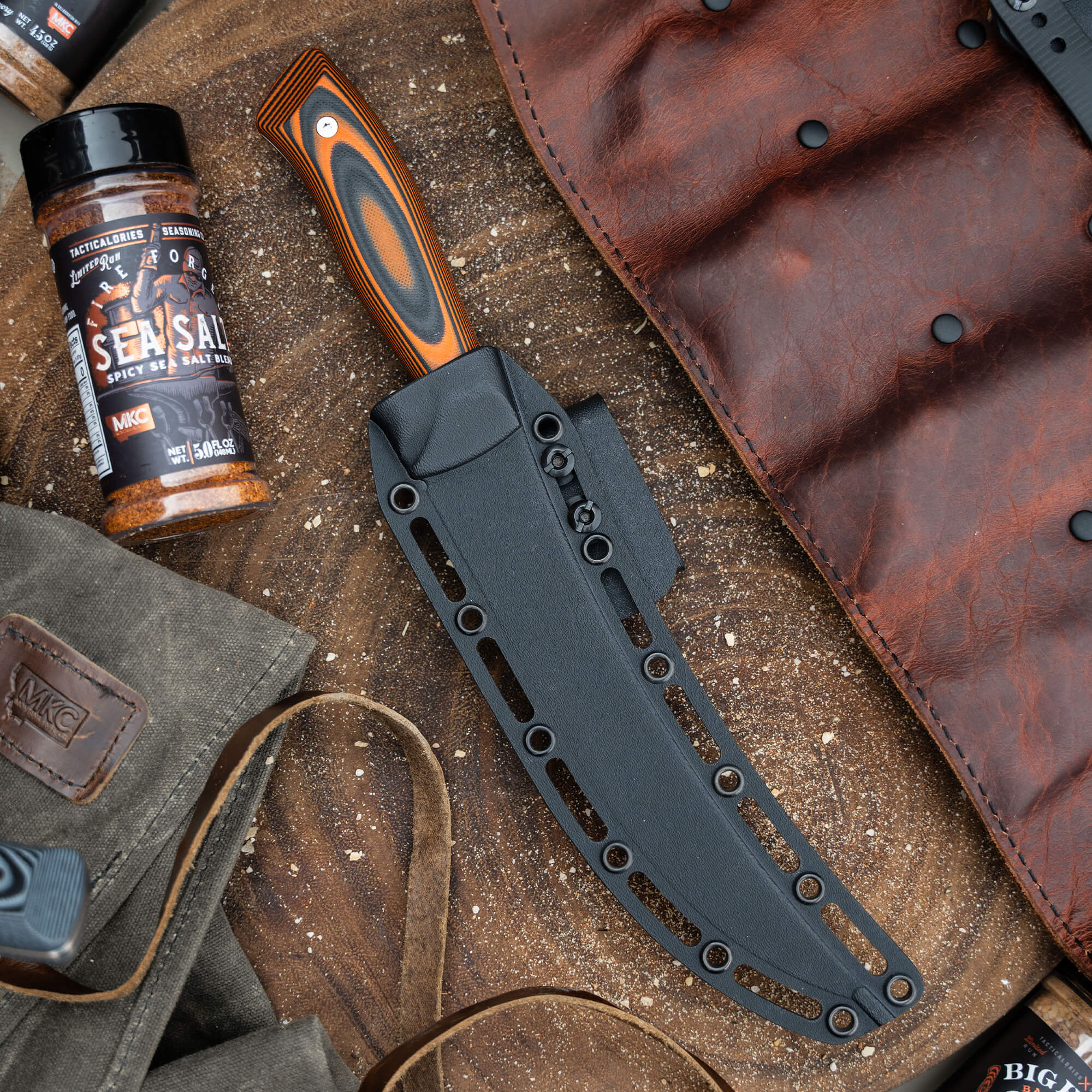 Alt text: "Premium quality breaking butcher knife with a wooden laminated orange and black handle in a black sheath, displayed next to a bottle of 'Sensai Spicy Sea Salt' seasoning and rugged leather accessories, illustrating an outdoor or rustic