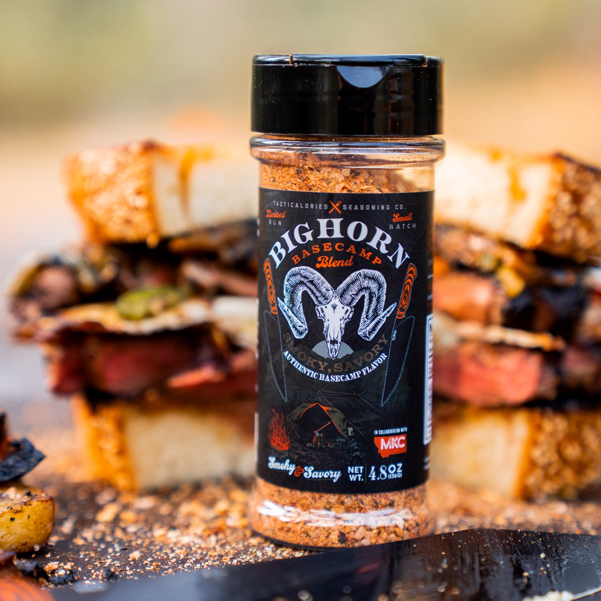 Alt text: "Bighorn Basecamp Seasoning blend in a clear container with a black lid, prominently displayed in front of a stack of grilled sandwiches and seasoned potatoes. The label features an illustration of a bighorn sheep and describes the seasoning