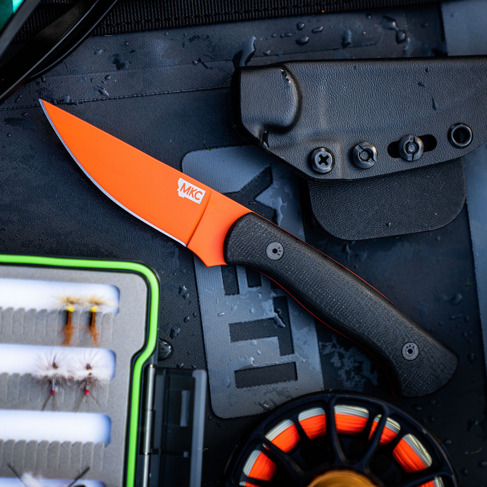 Blaze orange MKC Blackfoot 2.0 knife with black handle, positioned on a wet surface next to a black sheath and fishing accessories.